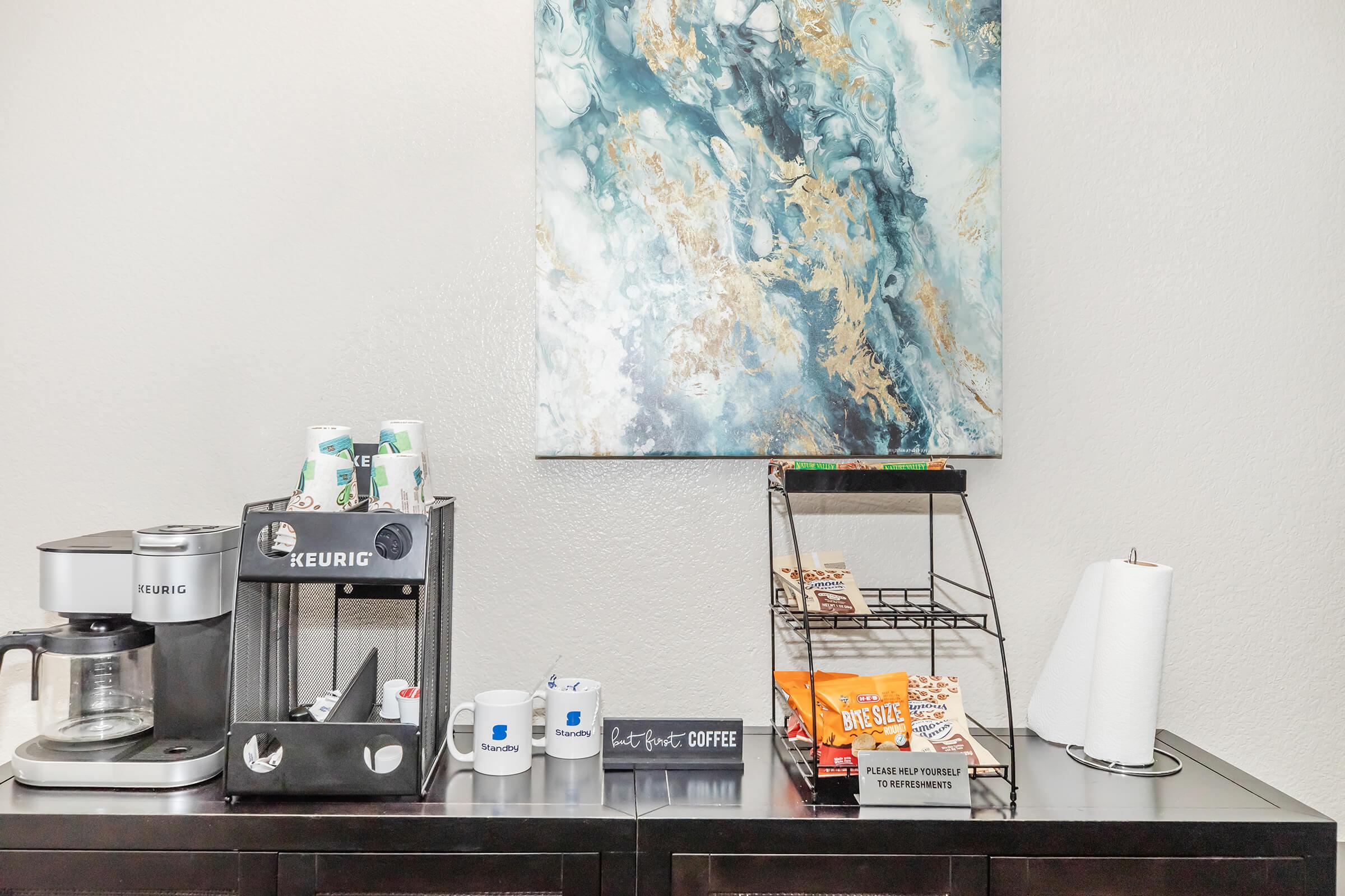 Coffee station featuring a Keurig machine, a coffee pot, and assorted cups. There are cans of beverage, a wire basket with snacks, and paper towels on a wooden surface. Above, an abstract art piece with blue and gold tones decorates the wall.