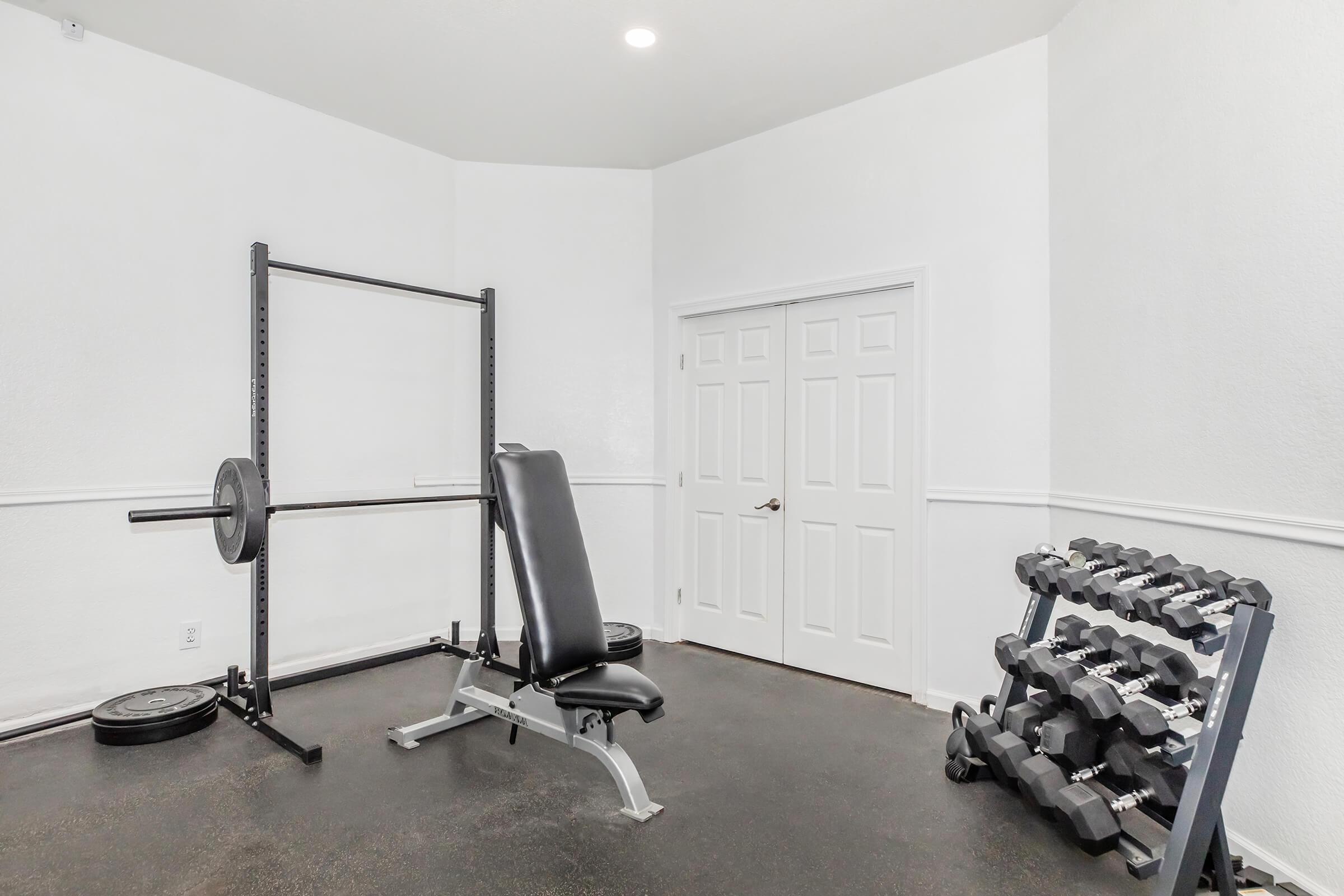 A spacious home gym featuring a weight bench with a barbell setup, a rack of dumbbells, and a plain white wall. The floor is dark, and there is a door on one side, creating a clean and minimalist workout environment.