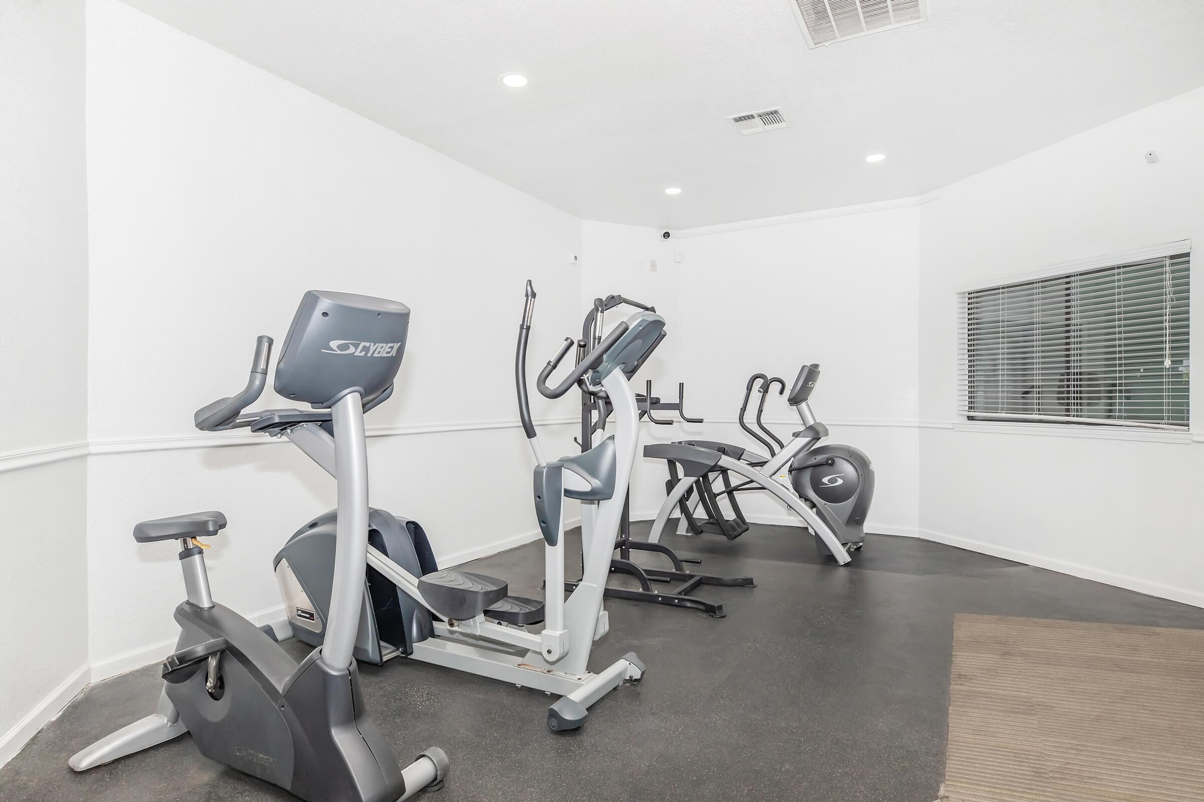 A well-lit fitness room featuring various exercise equipment, including a stationary bike, an elliptical machine, and a treadmill. The walls are painted white, and there is a large window allowing natural light to enter. The floor is covered with dark rubber flooring, providing a safe workout environment.