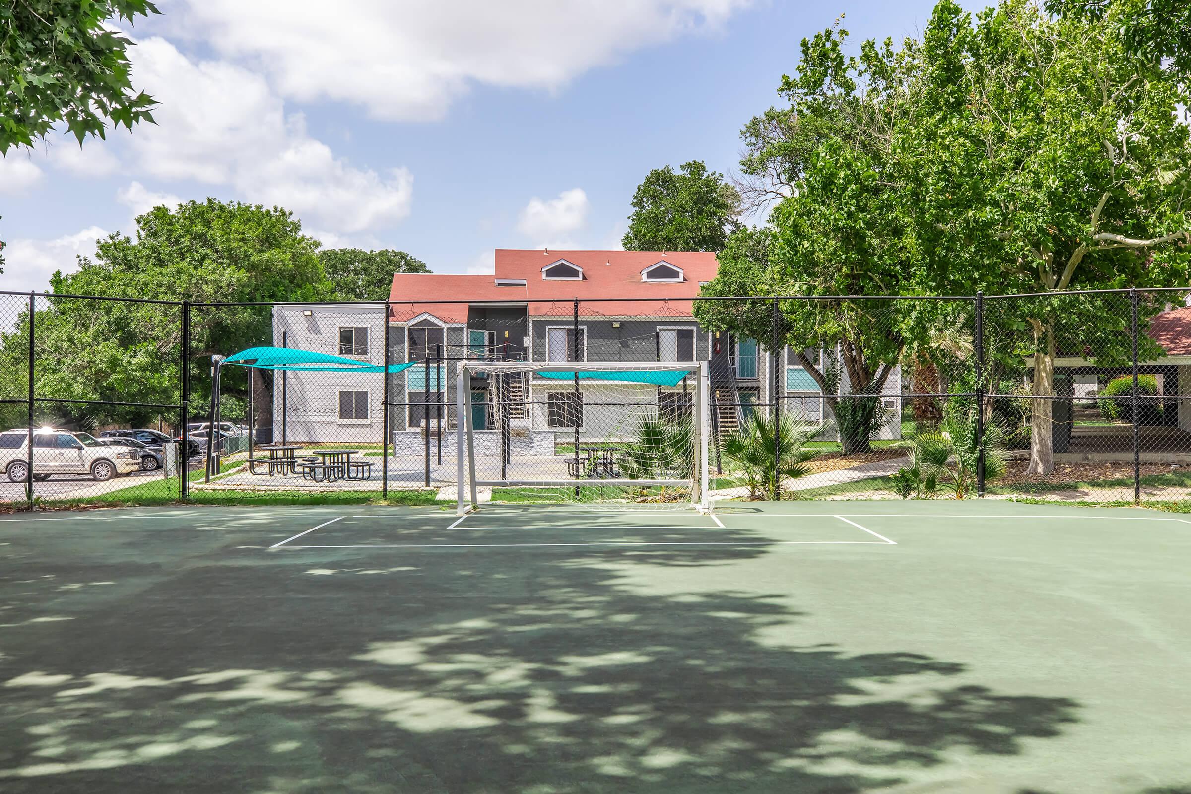 A fenced outdoor sports court with two soccer goals positioned in the foreground. In the background, a multi-story building with a red roof and several trees is visible. Blue canopies and picnic tables are set up nearby, creating a shaded area for relaxation. 