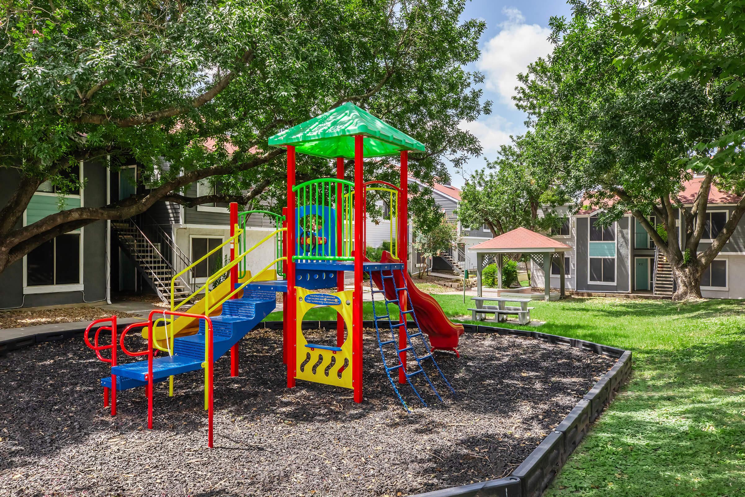 Colorful playground structure with slides and climbing features, surrounded by greenery and shaded by trees. The area is well-maintained, with mulch on the ground and nearby picnic tables, creating an inviting space for children to play.