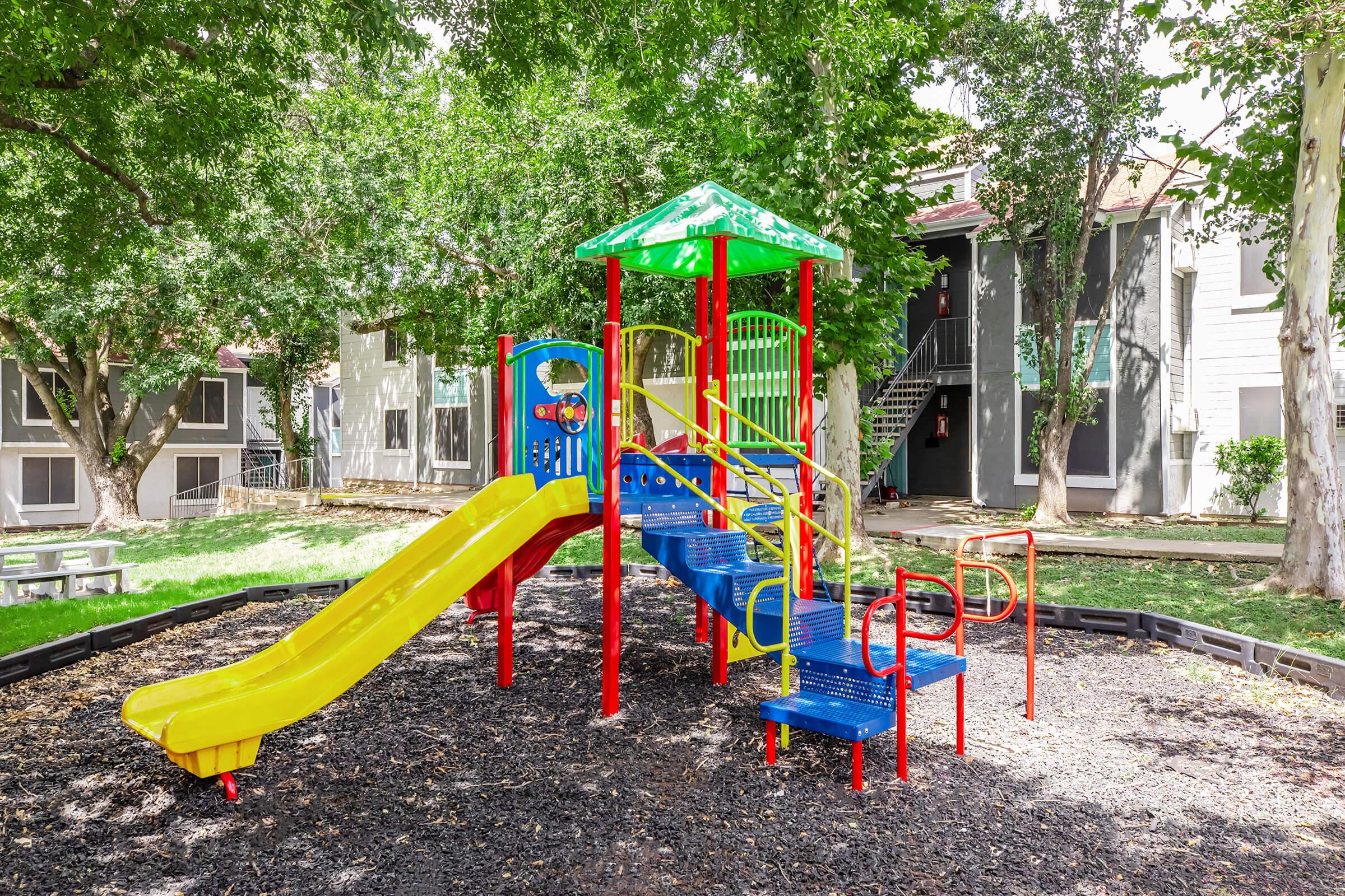 Colorful children's playground featuring a yellow slide, blue climbing steps, and a green roof. The area is surrounded by trees and grass, with an apartment building in the background. Safe, inviting space for kids to play and explore.