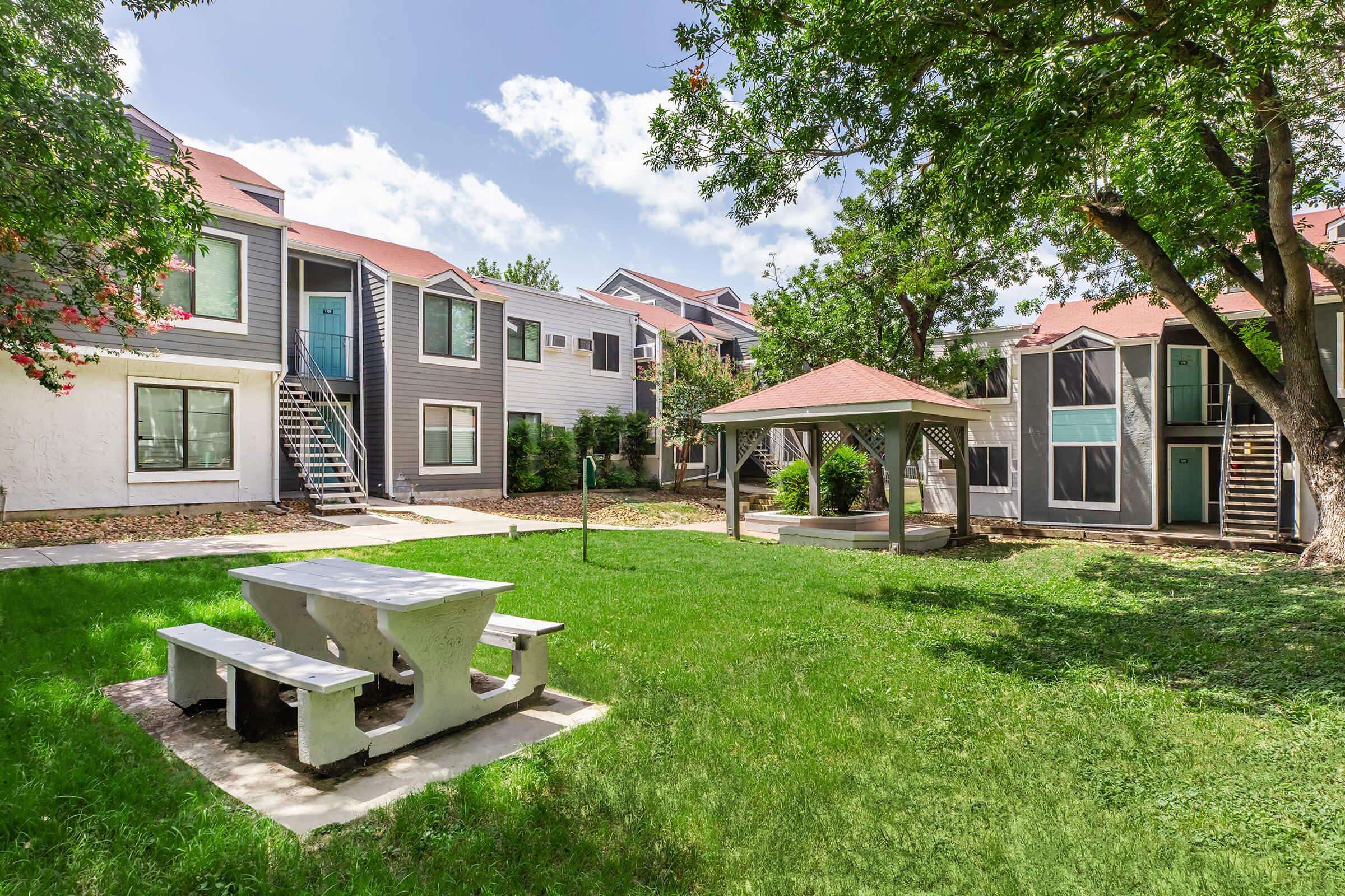 A residential area featuring green grass and a picnic table beneath a gazebo, surrounded by multi-story apartment buildings with varying exterior designs. The scene is bright with a partly cloudy sky and trees providing shade.