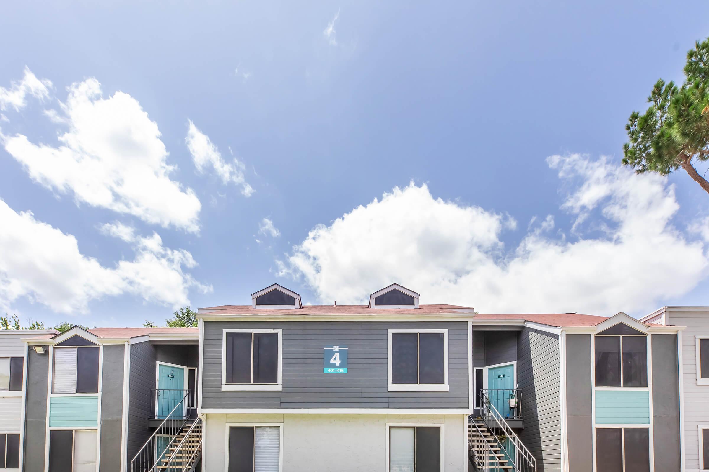 A two-story apartment building with a gray exterior, featuring blue doors and two small windows at the top. The number "4" is displayed prominently on the front. The sky is clear with scattered clouds, and there are trees in the background, creating a pleasant atmosphere.