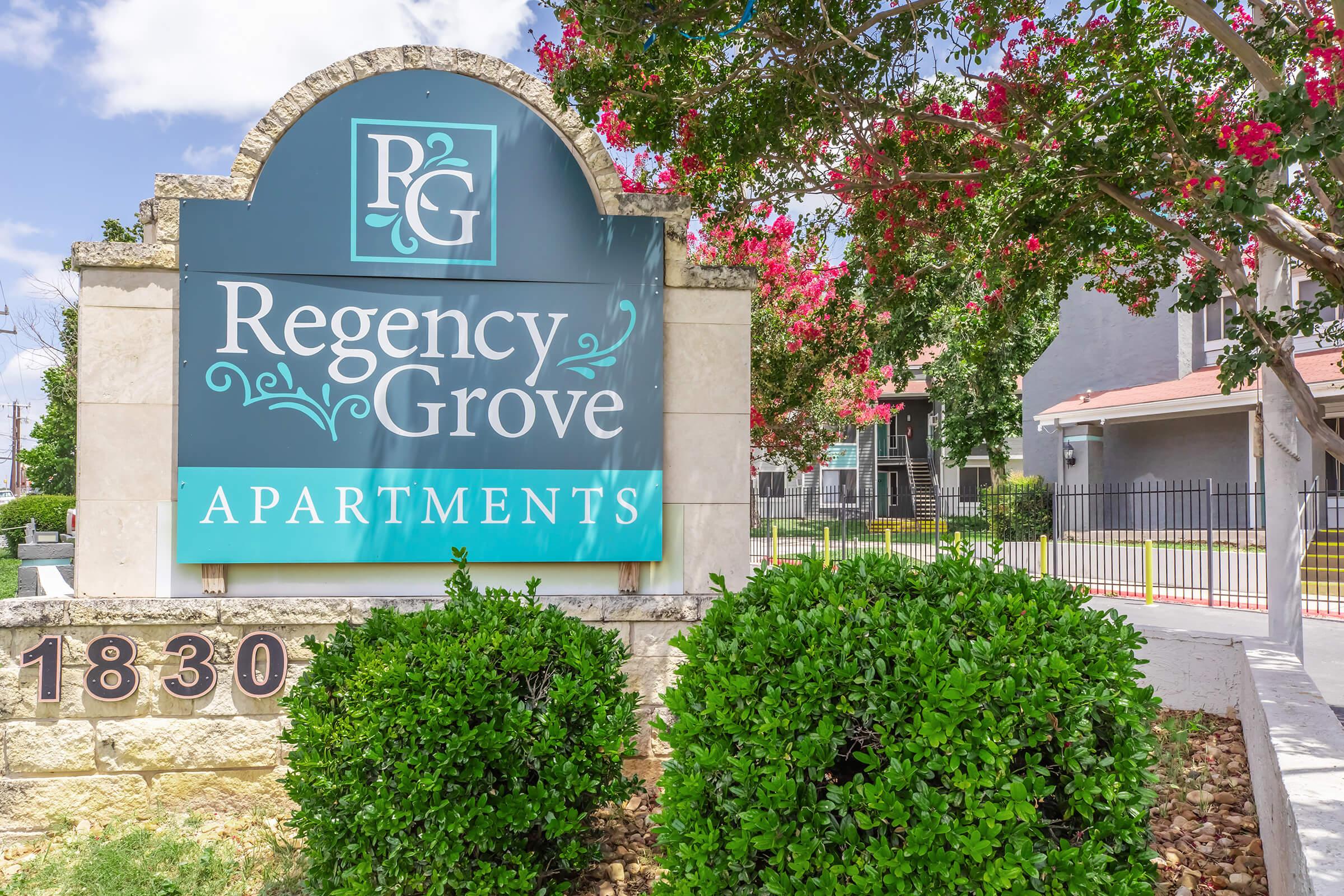 Sign for Regency Grove Apartments, featuring the name in decorative lettering, surrounded by lush green bushes and flowering trees. The building can be seen in the background, along with a clear sky. The address, 1830, is prominently displayed.