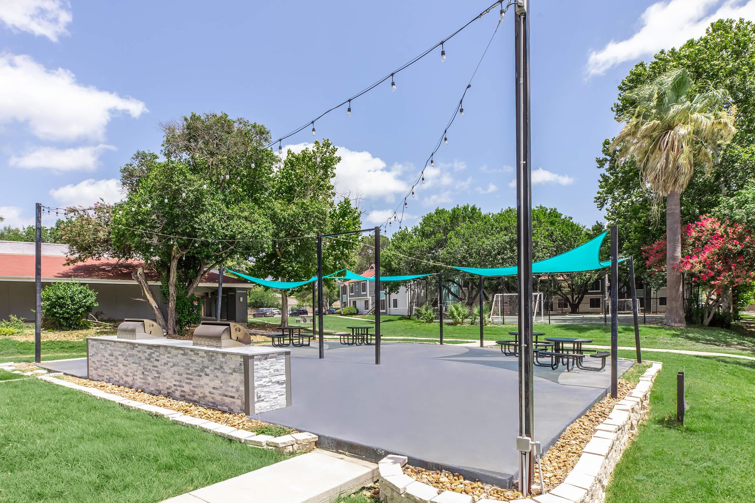 Outdoor community BBQ area with string lights, shaded by large fabric canopies. The space features a stone grill and several picnic tables on a concrete surface, surrounded by lush green grass and trees, creating a welcoming environment for gatherings and relaxation.