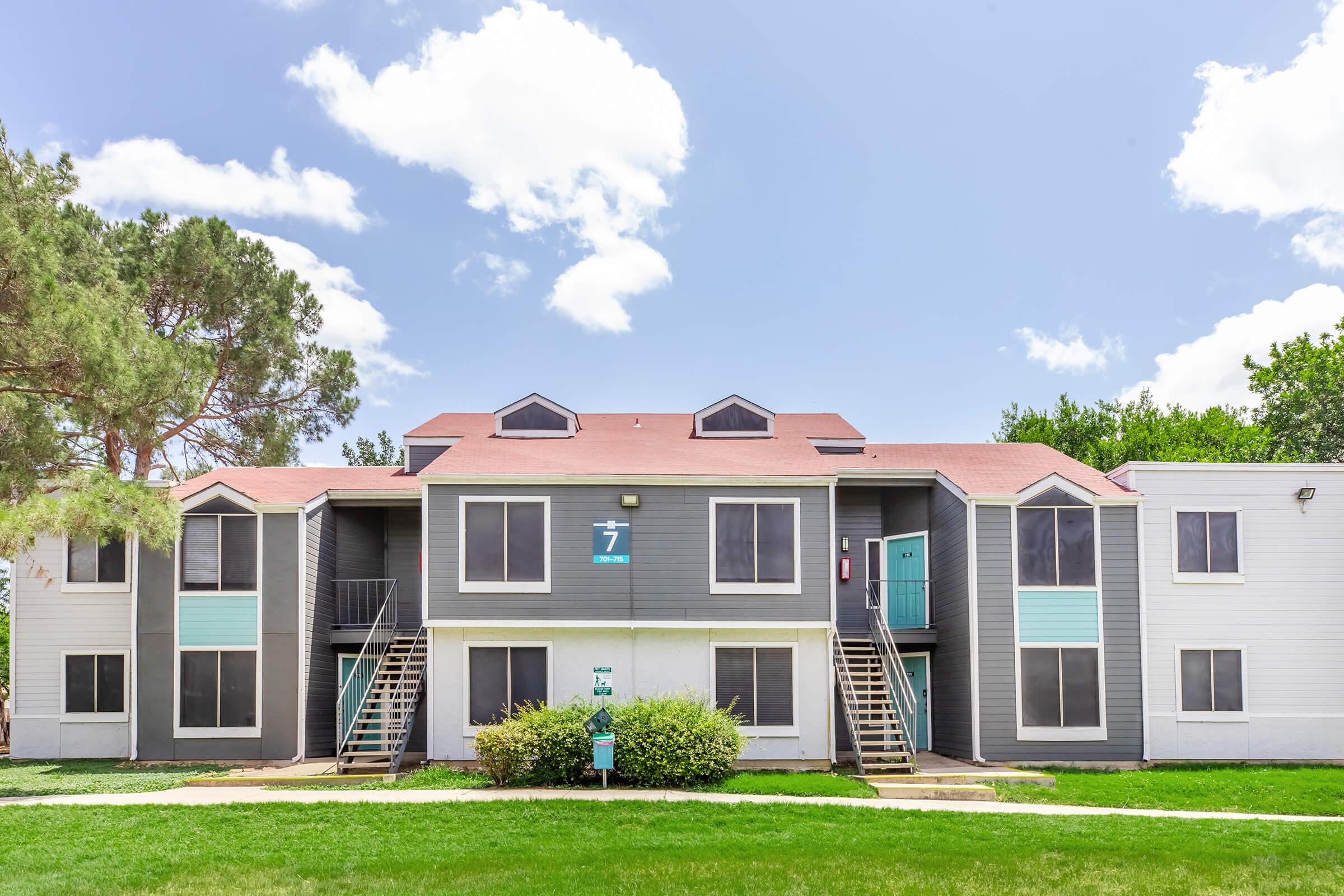 Two-story apartment building with multiple units, located among green grass and trees. The facade features a combination of dark gray and white siding, with prominent windows in each unit. Green landscaping surrounds the entrance, and a sign indicates the building number "7." The sky is clear with a few clouds.