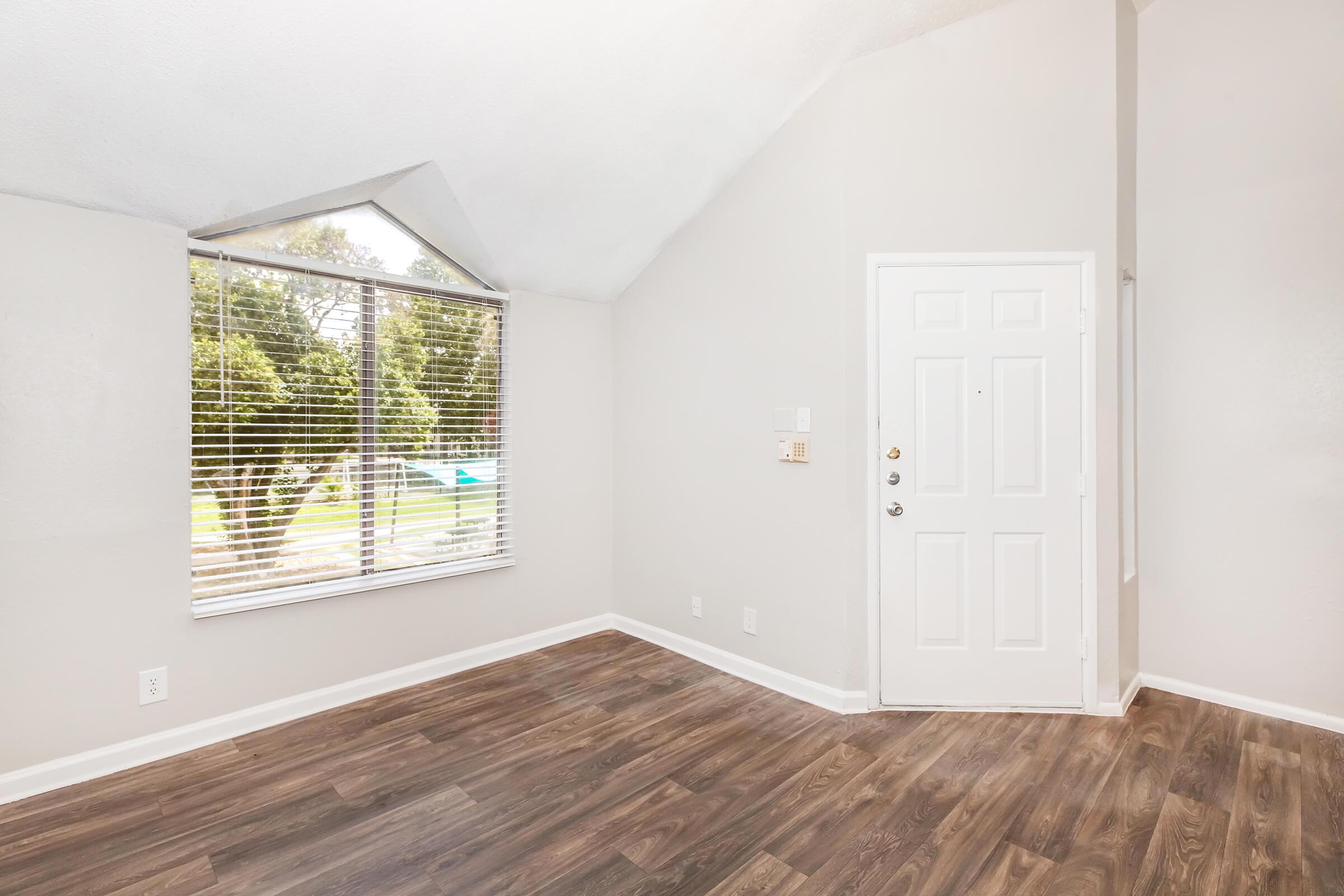 Empty room with a high ceiling and large window featuring blinds. The floor is covered in brown laminate, and there is a white door leading outside. The walls are light-colored, creating a bright and open feel. Natural light enters through the window, illuminating the space.