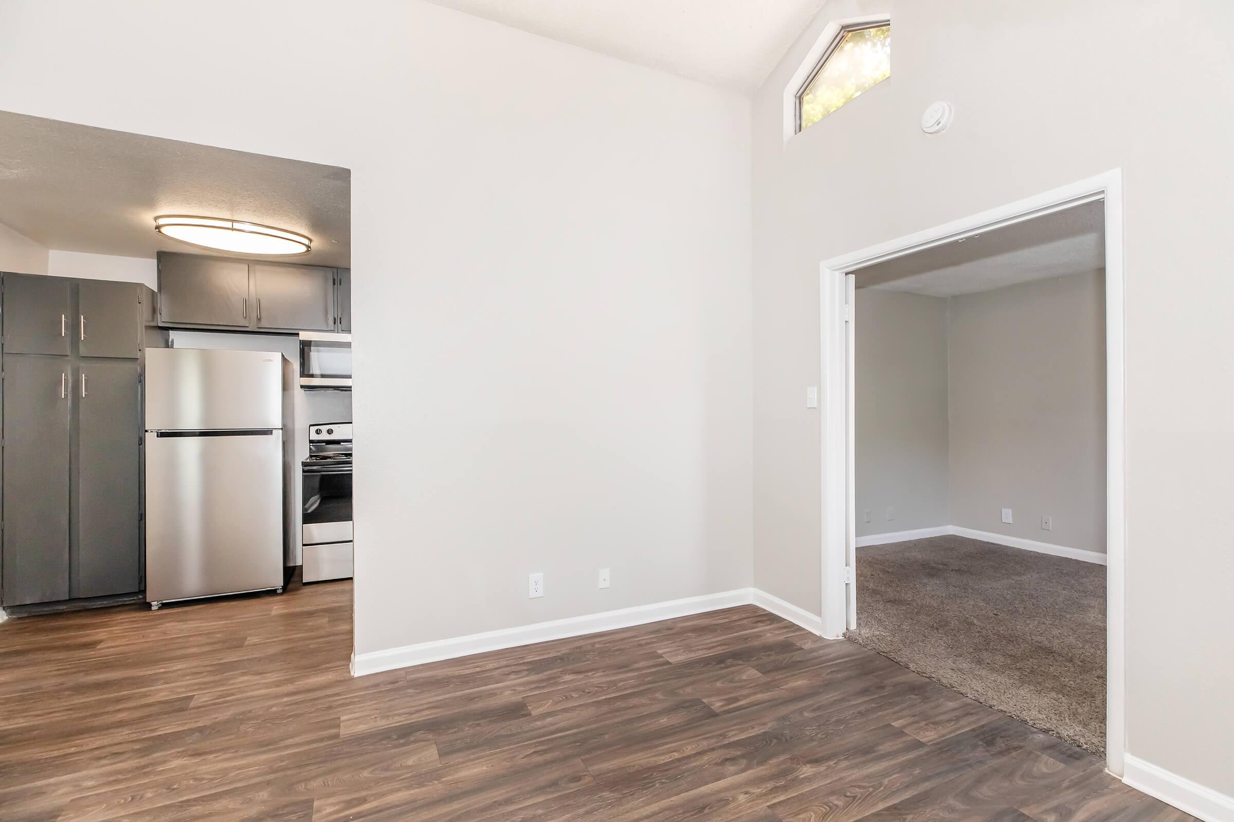 Interior view of an apartment featuring an open concept layout. On the left, a kitchen area with gray cabinets, stainless steel appliances, and a circular ceiling light. To the right, a living area with a carpeted surface and a doorway leading to another room, all painted in neutral tones. Natural light enters through a small window.