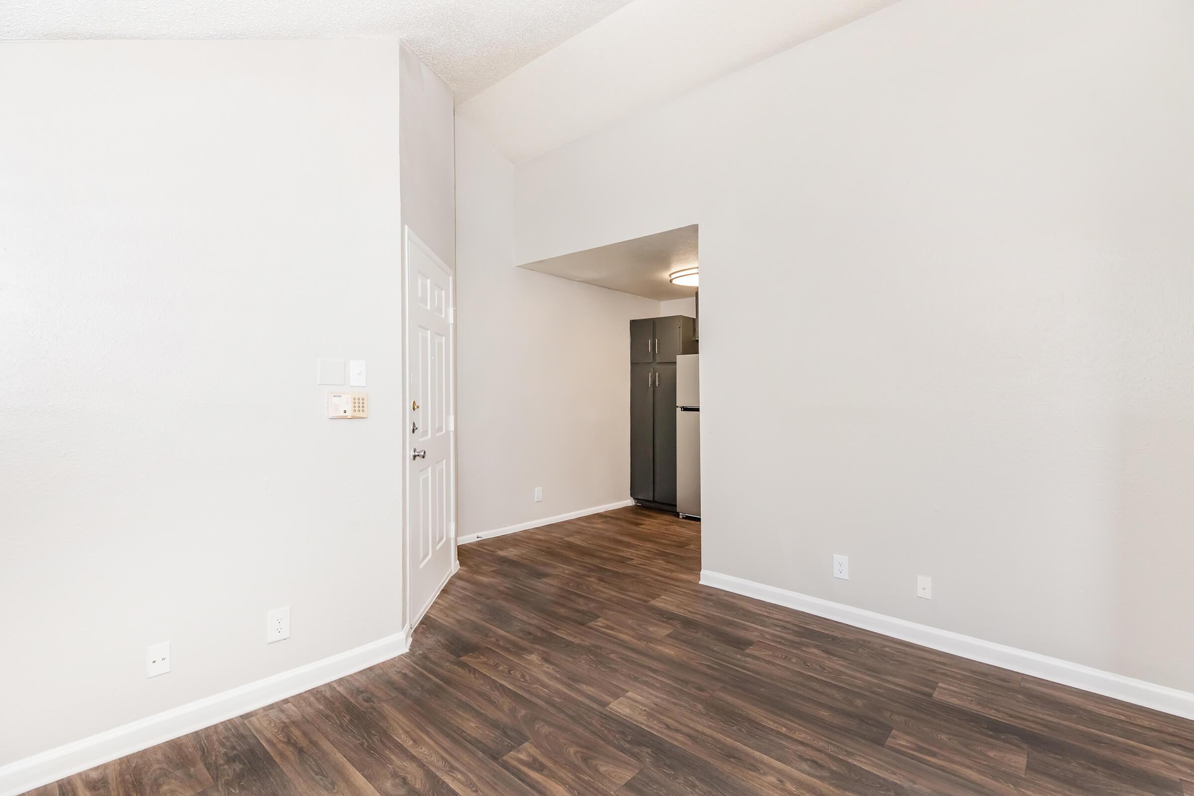 A spacious, vacant room with light-colored walls and a hardwood floor. A doorway leads to a kitchen area, partially visible with a refrigerator. Natural light enhances the clean, uncluttered look of the space, creating an inviting atmosphere suitable for various furnishings or decorations.