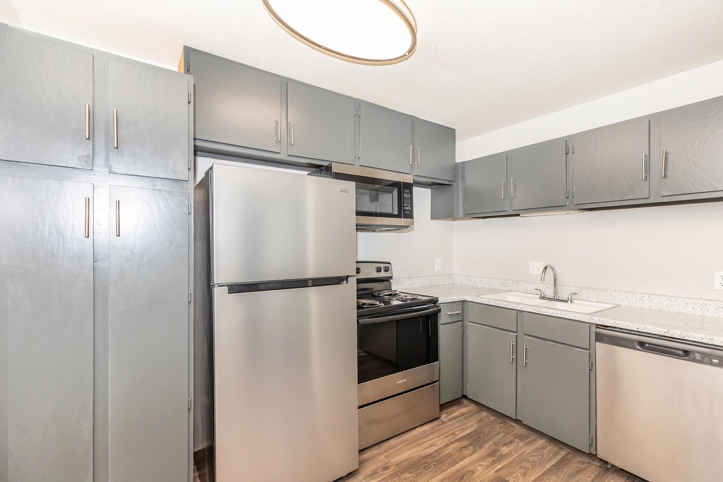 Modern kitchen featuring gray cabinetry, stainless steel refrigerator, and appliances including an oven and microwave. The countertop is light-colored granite, and the flooring is dark wood, creating a contemporary aesthetic. Natural light possibly coming from an overhead fixture.
