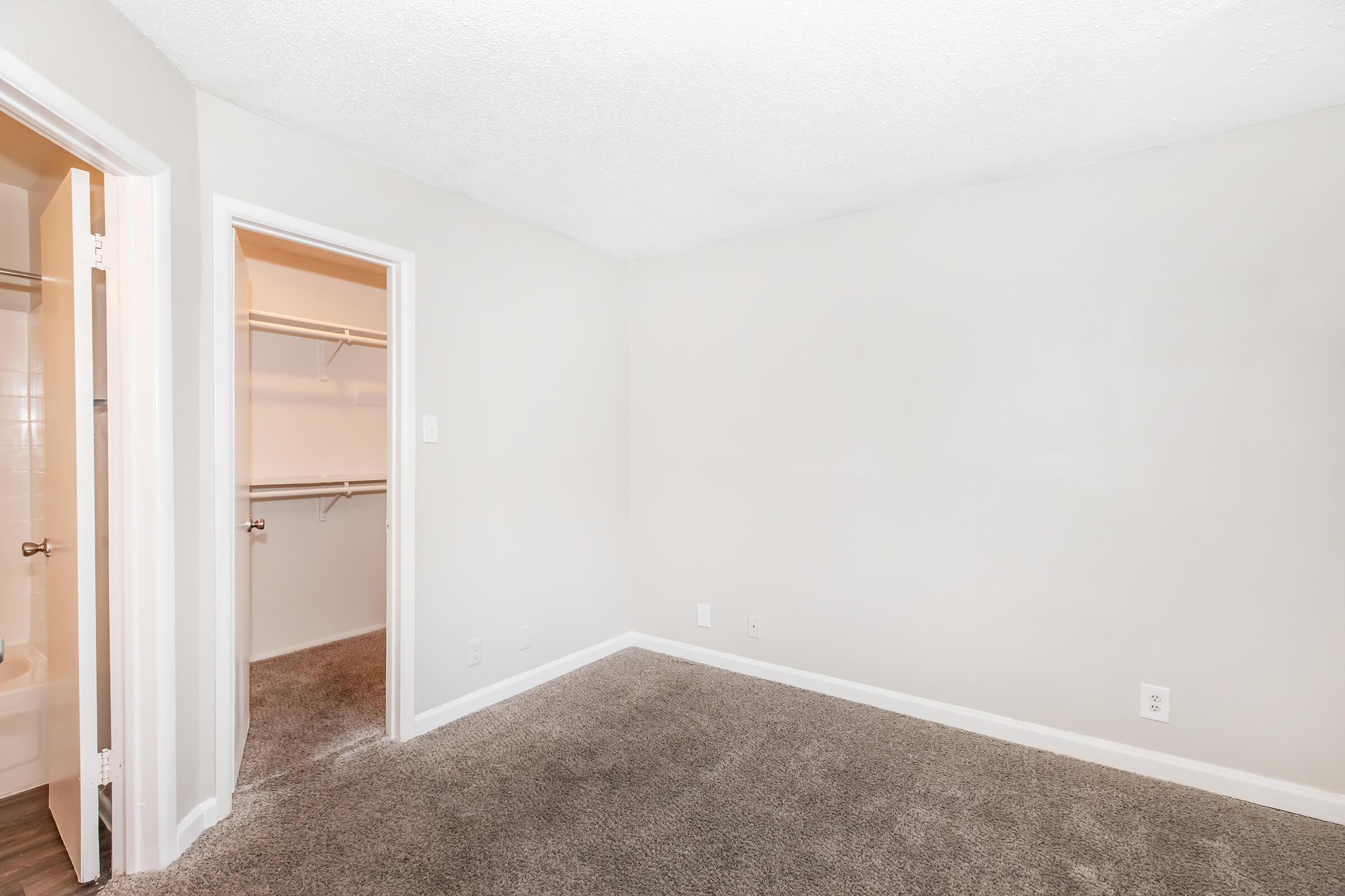 A spacious, empty room with light gray walls and a carpeted floor. A doorway on the left leads to a closet space, while another doorway on the right opens to a bathroom. The room is well-lit, featuring a clean and simple aesthetic.