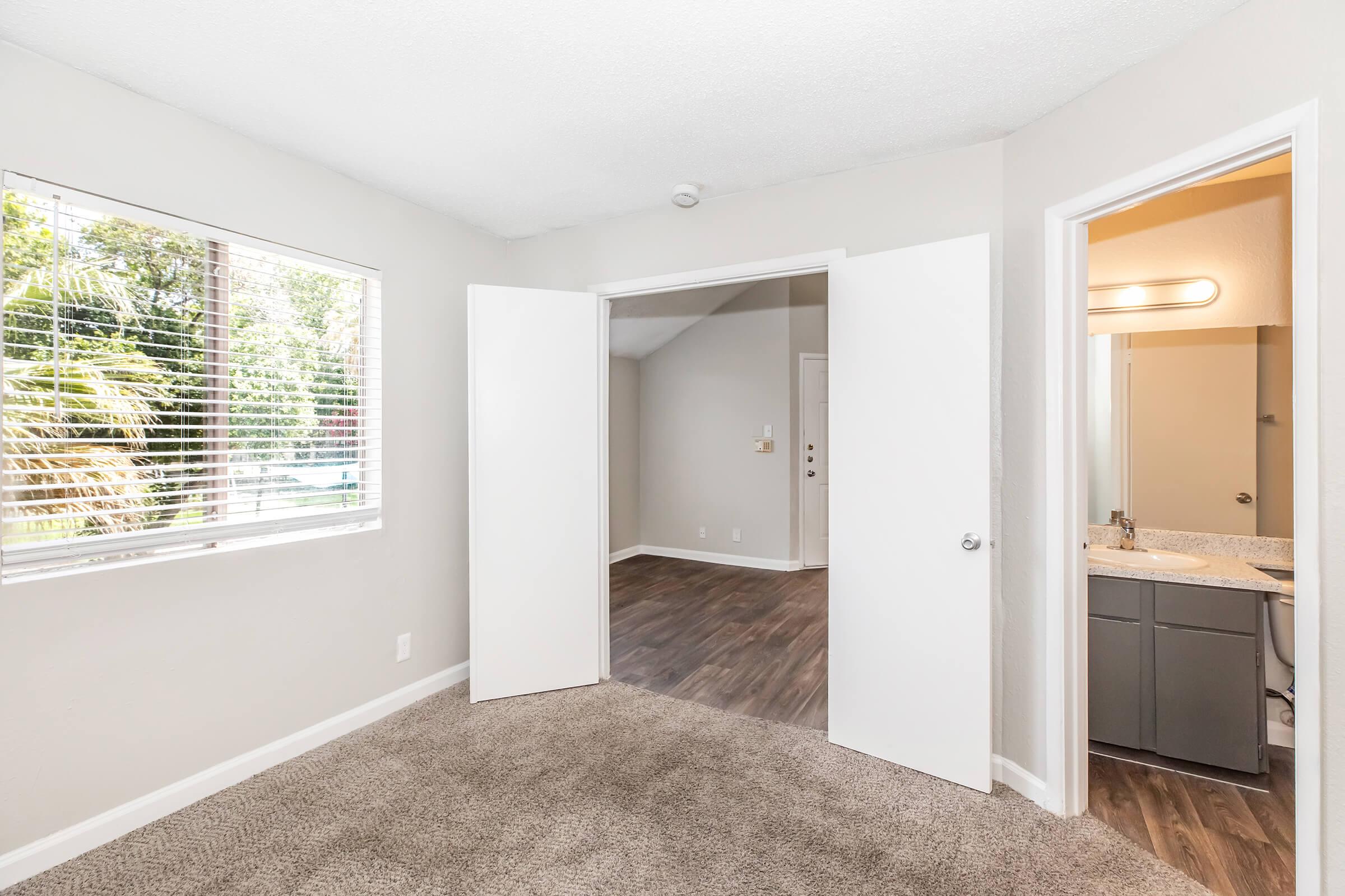A bright, airy room with light-colored walls and carpeted flooring. Large windows allow natural light to flow in, showcasing views of greenery outside. Two doors lead to other spaces: one to a bathroom with modern fixtures and the other to an adjoining room with a spacious feel.