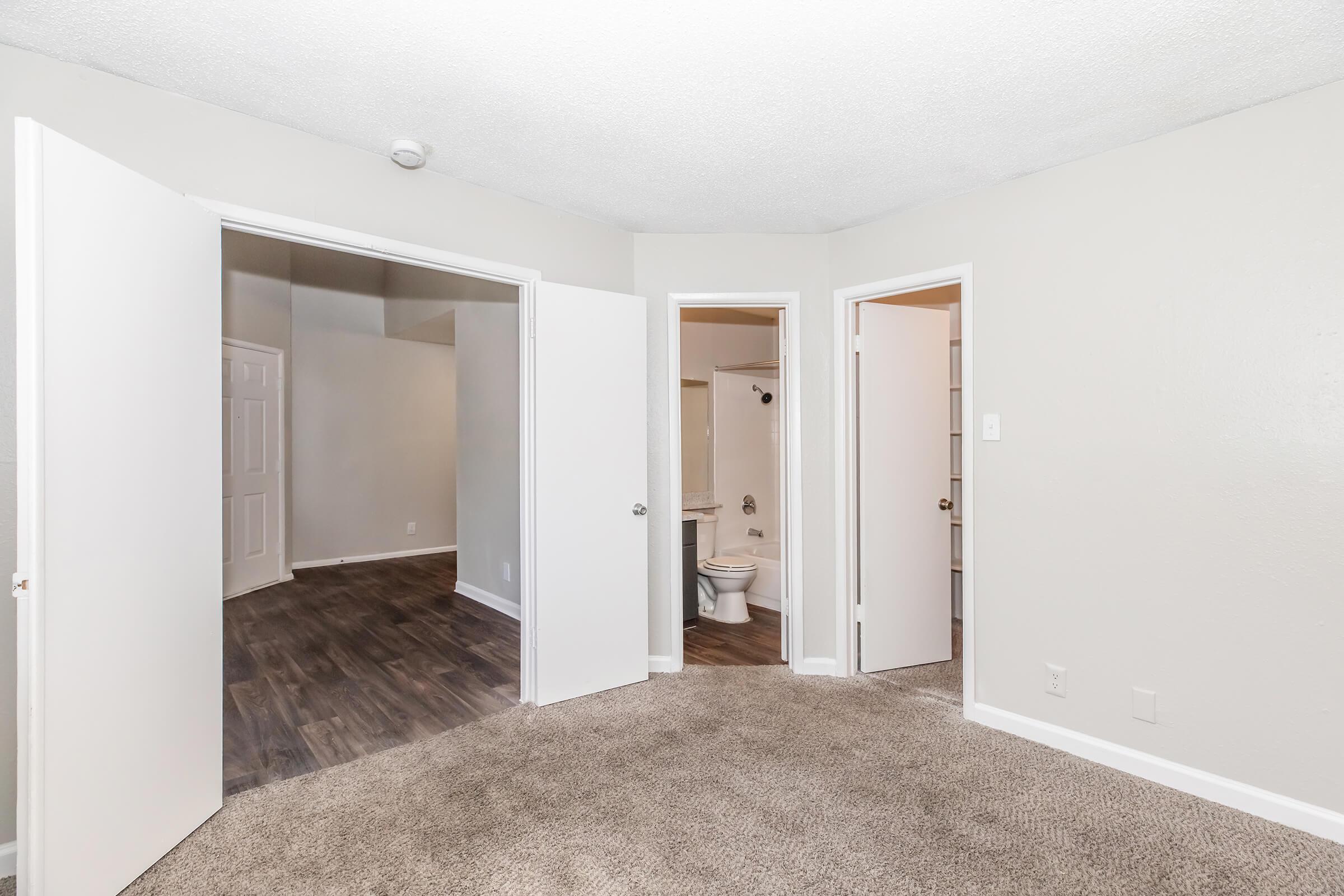 A well-lit room featuring beige carpet and light gray walls. Two white doors lead to adjacent spaces, one opening to a bathroom and the other to another room. The room is partially empty, with no furniture visible, creating a spacious and inviting atmosphere.