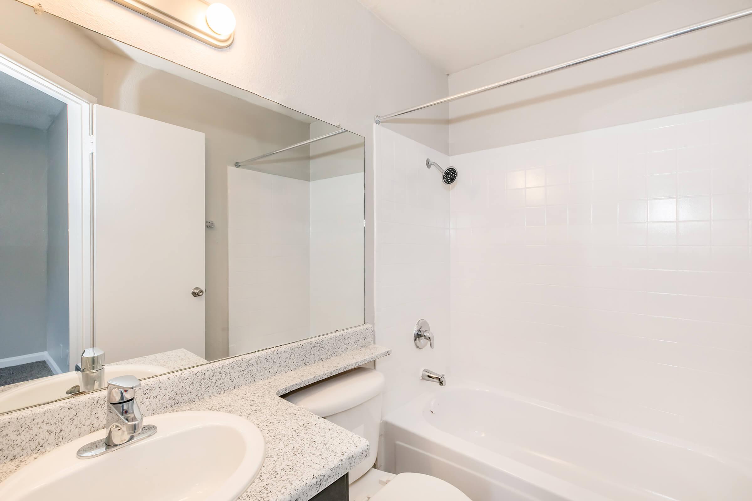 A clean bathroom featuring a white tiled shower and bathtub, a mirrored vanity with a sink, and light-colored walls. The scene includes a door leading to another room and gray carpeting visible in part of the image.