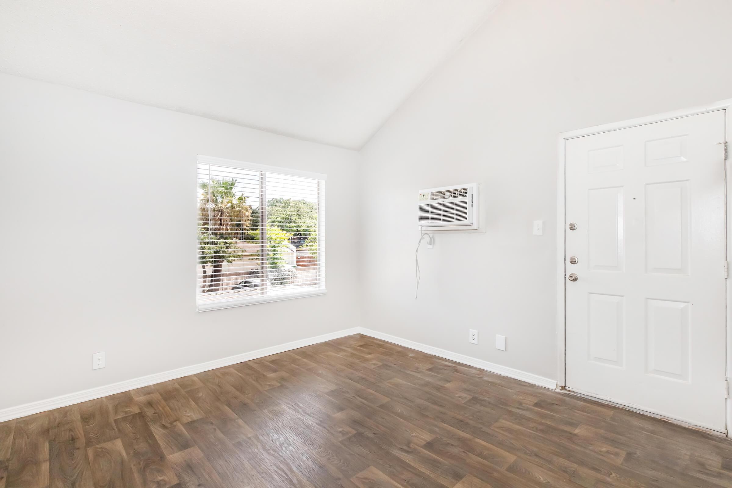 A bright, empty room with light gray walls and wood-style flooring. There's a large window with blinds letting in natural light, and an air conditioning unit above it. A white door with a doorknob is visible on the right side. The space is clean and uncluttered, suitable for various uses.