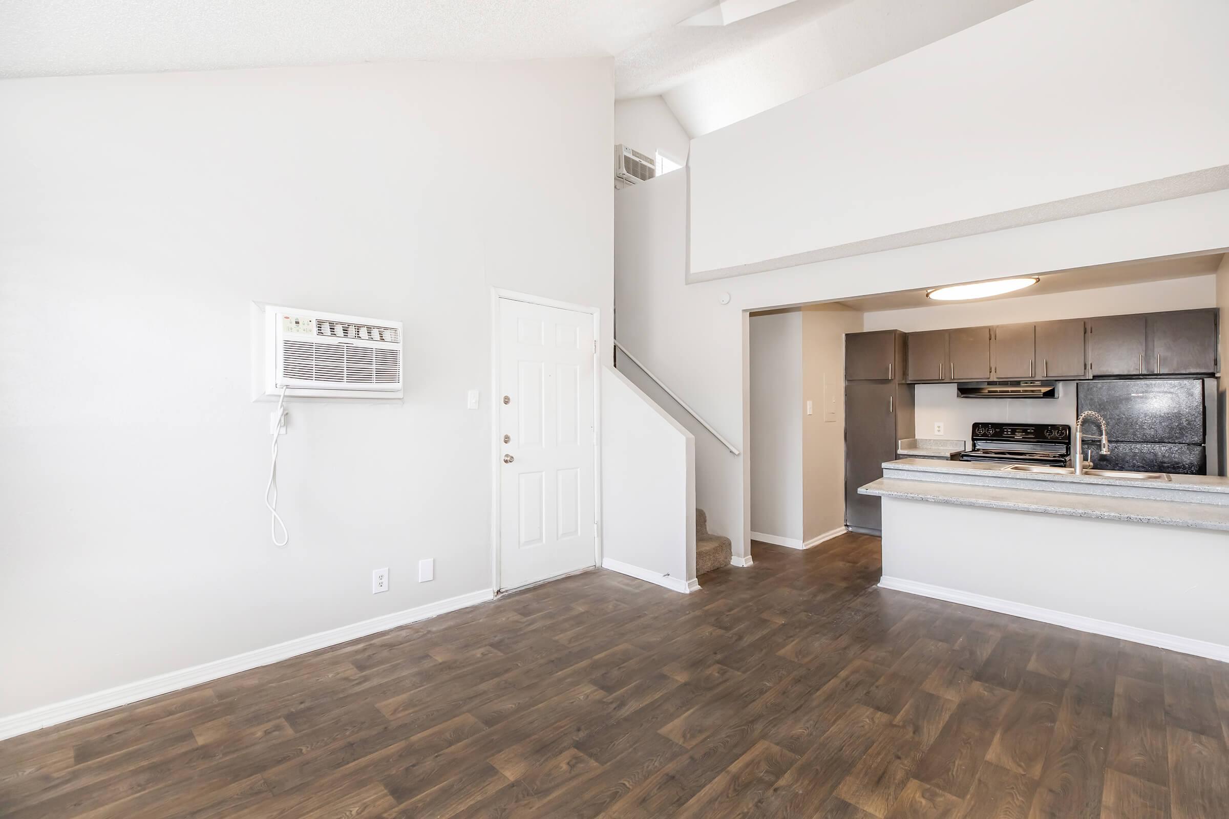 Bright and spacious interior of an apartment featuring a living area with wood-like flooring, an air conditioning unit on the wall, and a staircase leading to an upper floor. The kitchen, visible in the background, has dark cabinets and a countertop, enhancing the modern look of the space.