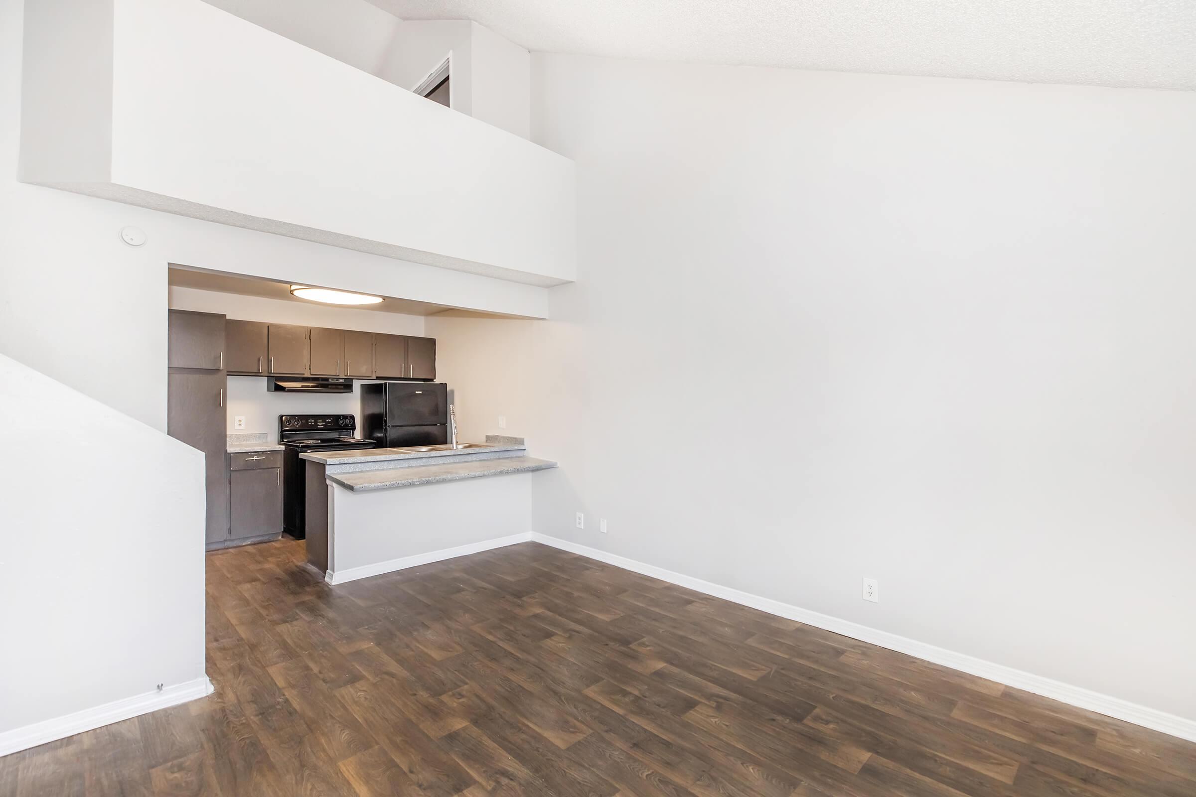 Interior of a spacious, modern living area showcasing hardwood floors, a high ceiling, and an open layout. The kitchen area features dark cabinets and appliances, partially visible through an archway. Bright, neutral walls enhance the room's airy feel, with natural light filtering in from above.