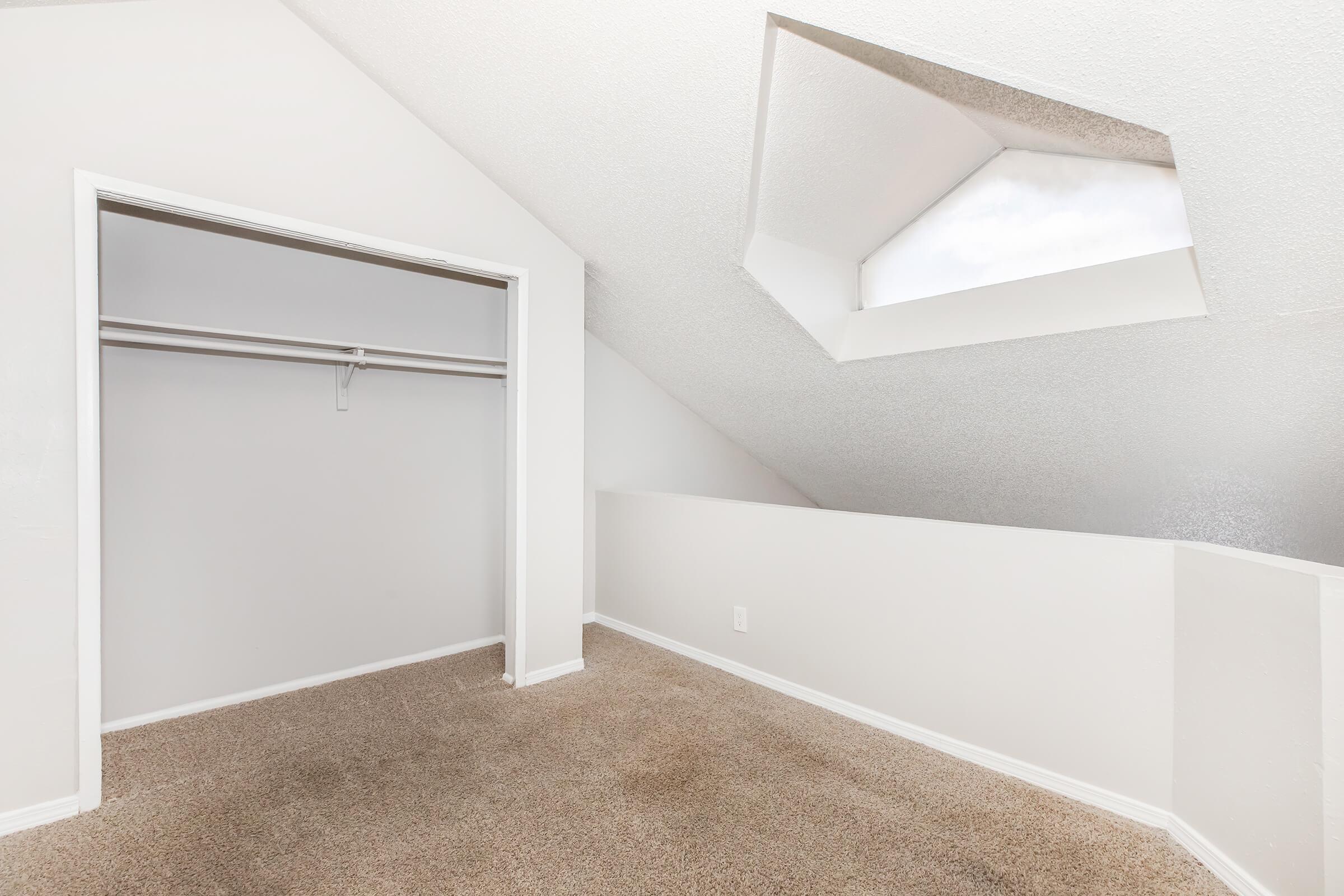 A bright, empty room with sloped ceilings and light-colored walls. There's a small closet on one side and a unique skylight that adds natural light. The carpet is beige, creating a warm atmosphere, and the overall space is clean and modern. Ideal for a study or additional storage.