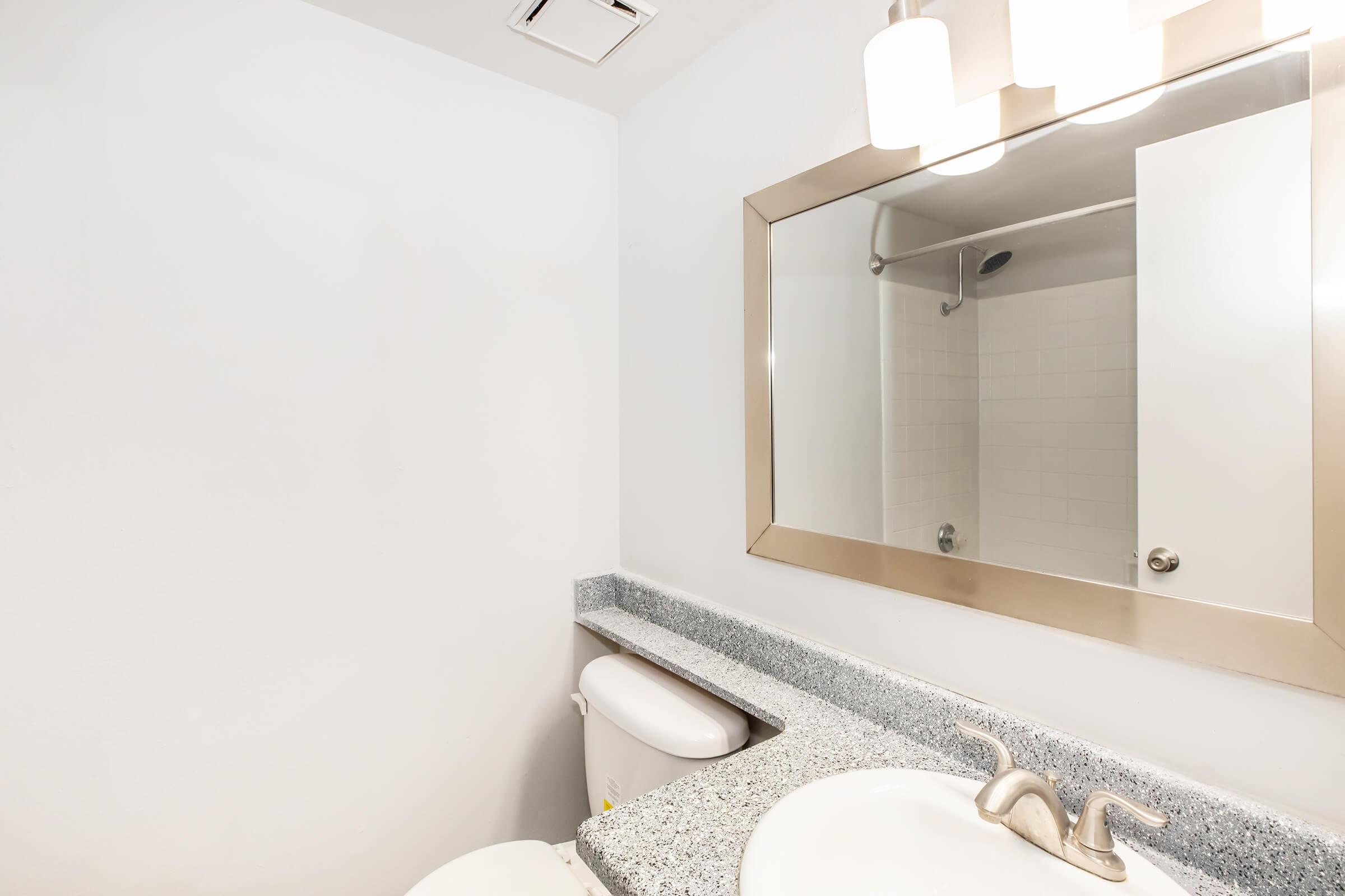 A clean, modern bathroom featuring a large mirror with a silver frame, a granite countertops, a toilet, and a shower area with transparent doors. The walls are painted white, and there is overhead lighting from a three-bulb fixture.