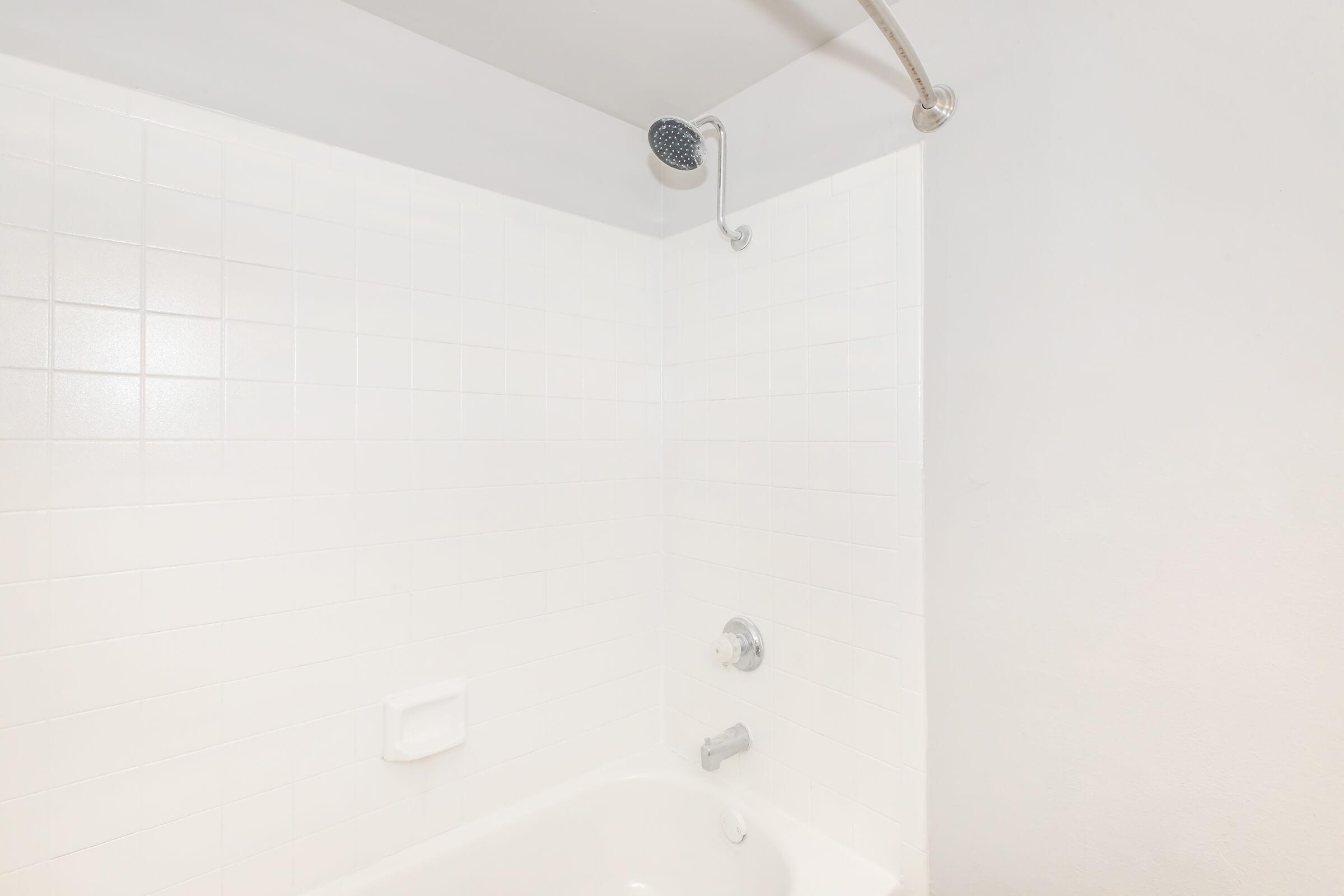 A clean, modern shower area featuring white tiled walls and a white bathtub. The showerhead is mounted on the wall, above the bathtub, with a simple chrome faucet and control. The design is minimalistic, giving a fresh and airy appearance to the space.