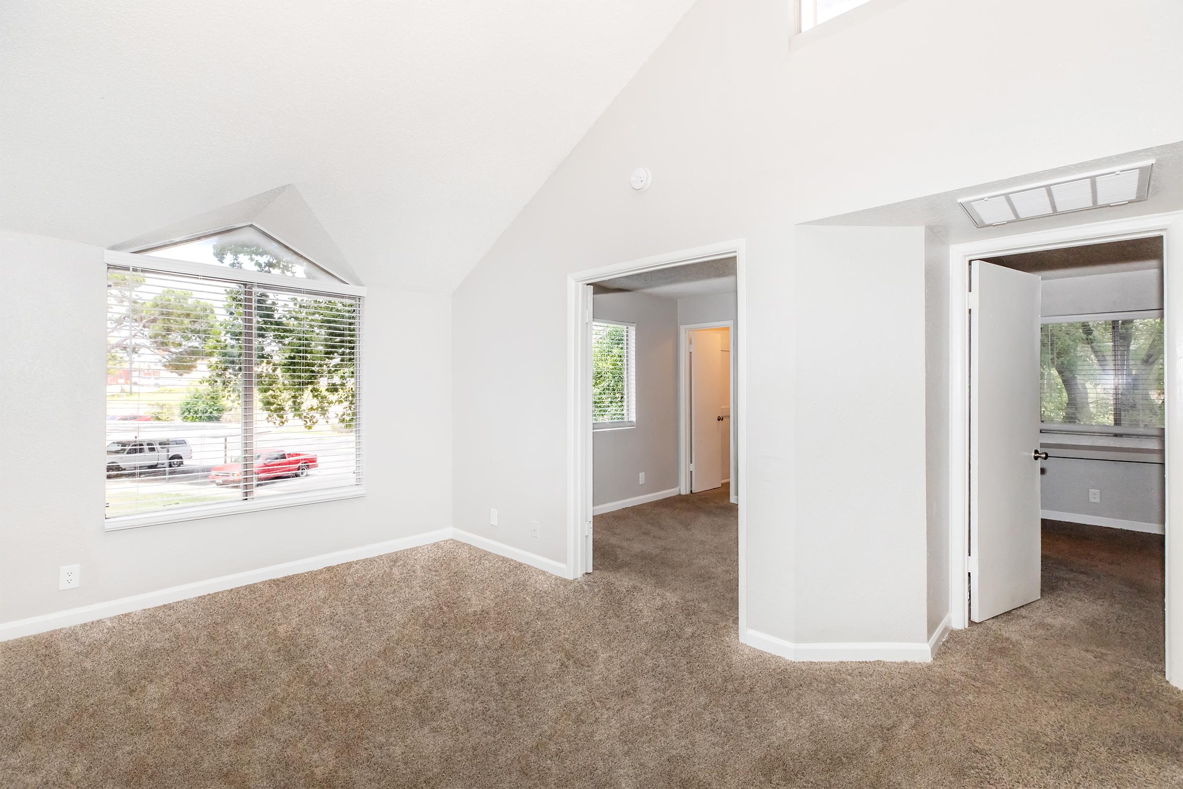 A spacious, well-lit room featuring high ceilings and multiple windows with shades. The walls are painted in a light color, enhancing the brightness. The floor is covered in plush carpet. Doorways lead to adjoining spaces, creating an inviting atmosphere suitable for a living area or bedroom.