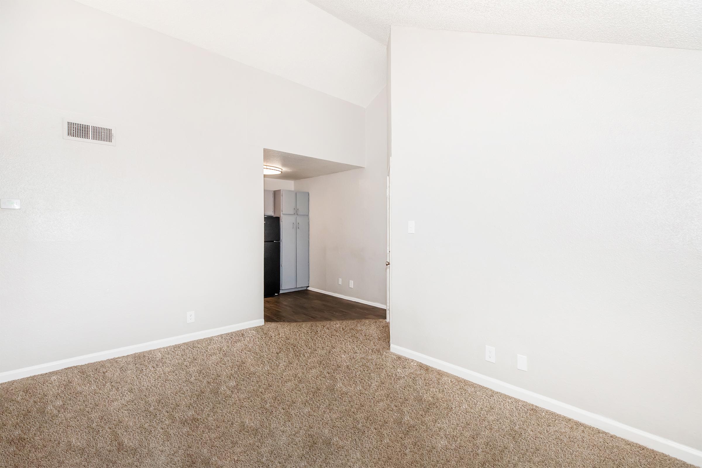 A spacious, light-colored room with carpet flooring, featuring a high ceiling. On the left, there's a doorway leading to another room, and in the background, a glimpse of a kitchen area with a refrigerator is visible. The walls are painted in a neutral tone, creating an open and airy feel.