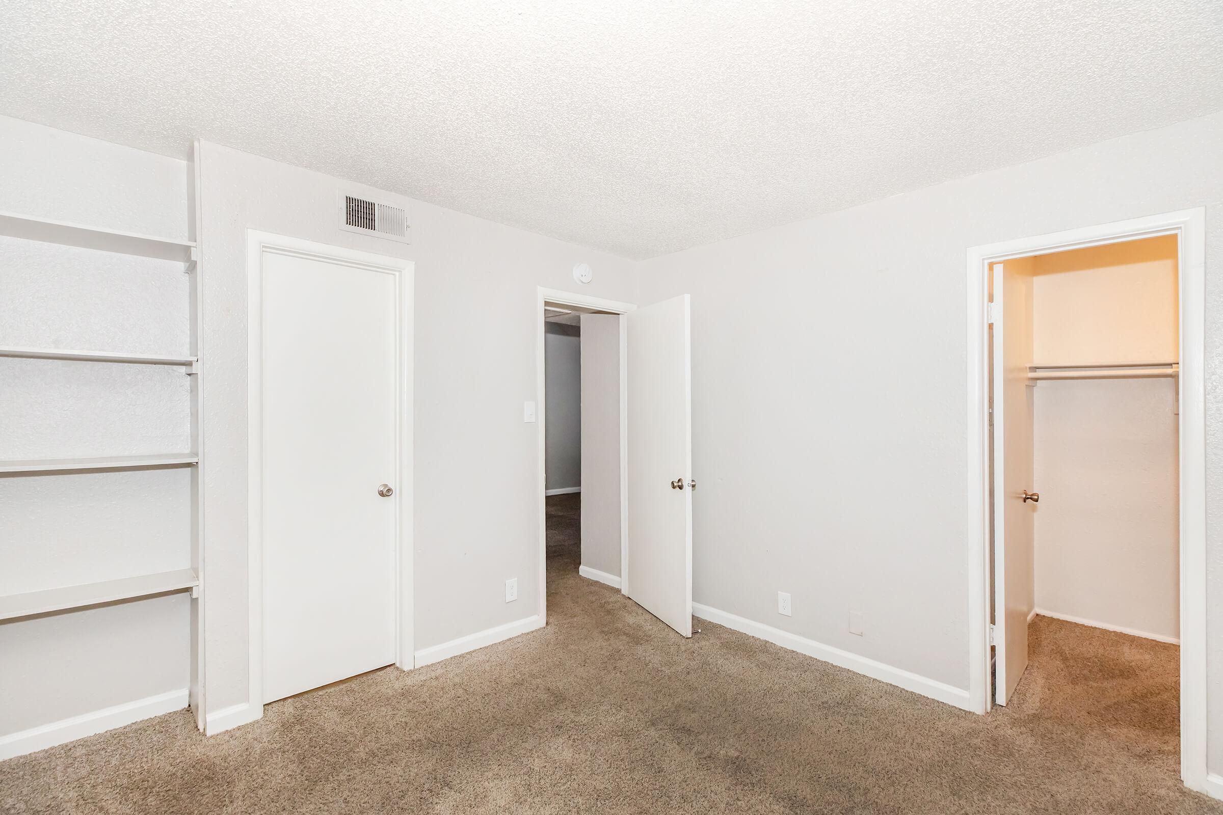 A neutral-colored room with carpeted flooring, featuring two closed doors, one leading to a closet and the other to another space. There is also a built-in shelf along one wall, providing storage options. The walls are painted light gray, creating a spacious and inviting atmosphere.