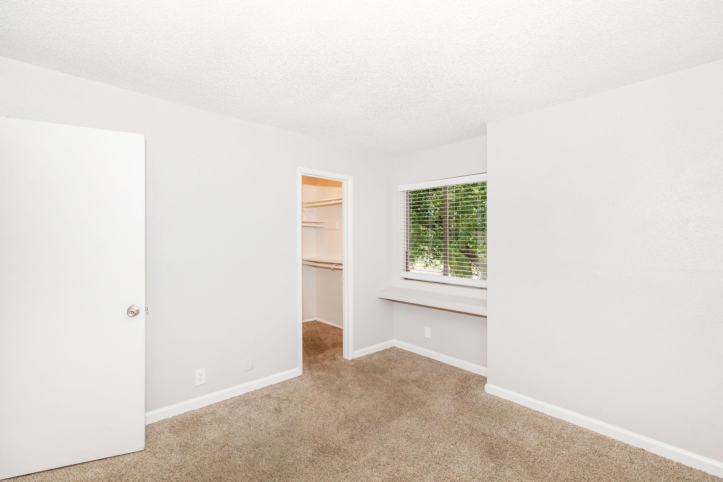 A small, empty room with light gray walls and beige carpet. It features a window with blinds, a closet with a single door, and a built-in shelf under the window. The lighting is bright, providing a clean and airy feel to the space.