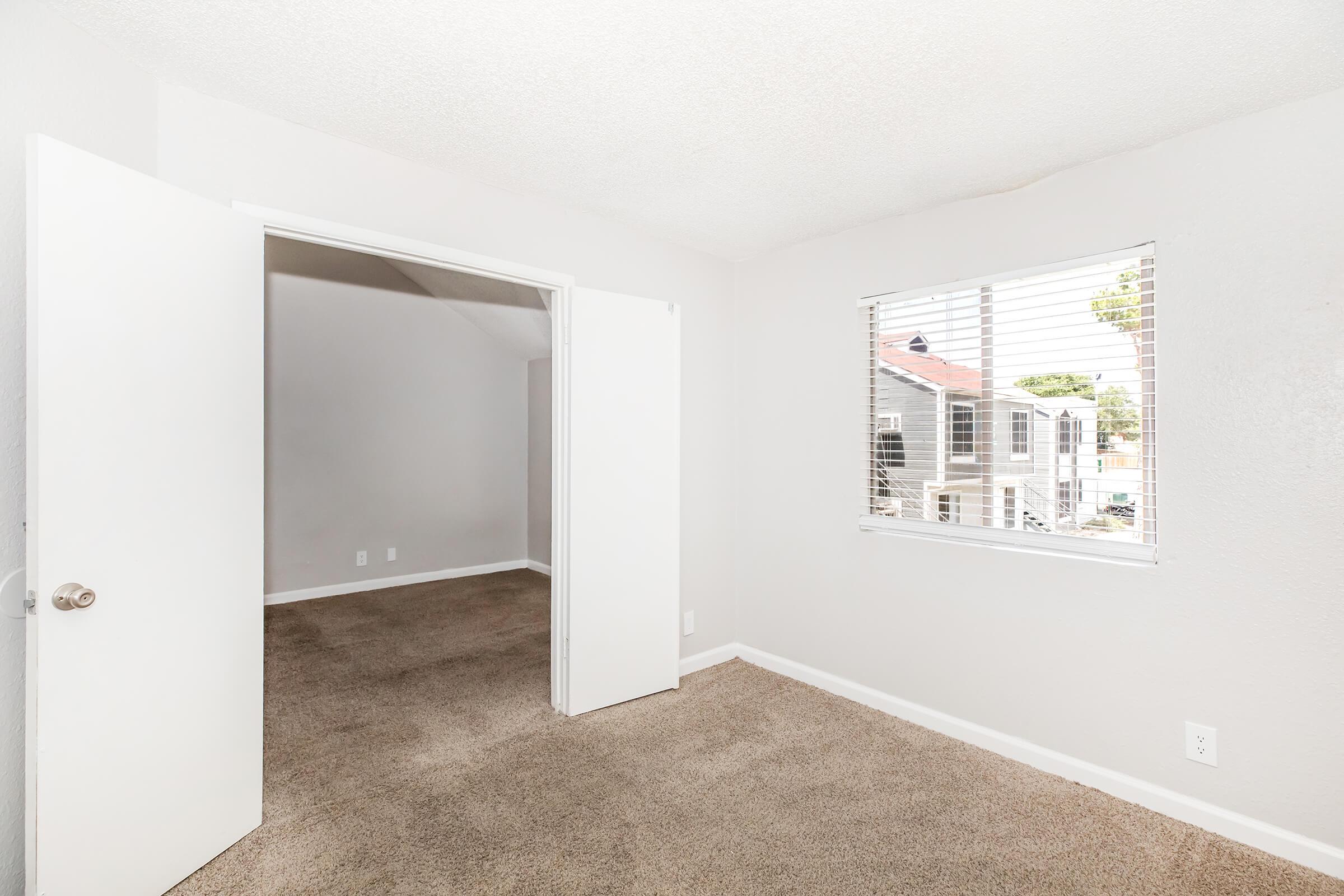 A bright, empty room with light beige carpet and gray walls. Two open doors lead to an adjacent room, and there is a window on the right side with blinds partially drawn. Natural light enters through the window, illuminating the space. Standard light fixtures are visible on the ceiling.