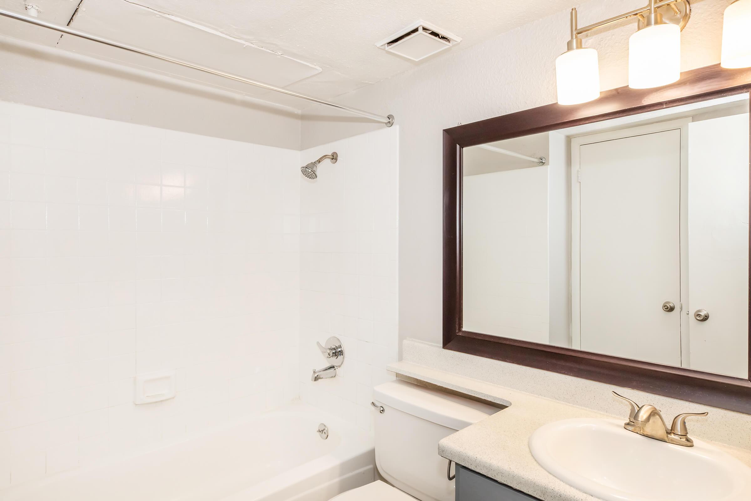 A clean, modern bathroom featuring a white bathtub, a toilet, and a sink with a countertop. The space includes a large mirror with three light fixtures above it. The walls are plain white, and there is a silver showerhead and a towel bar on the wall. A door leads to another room.