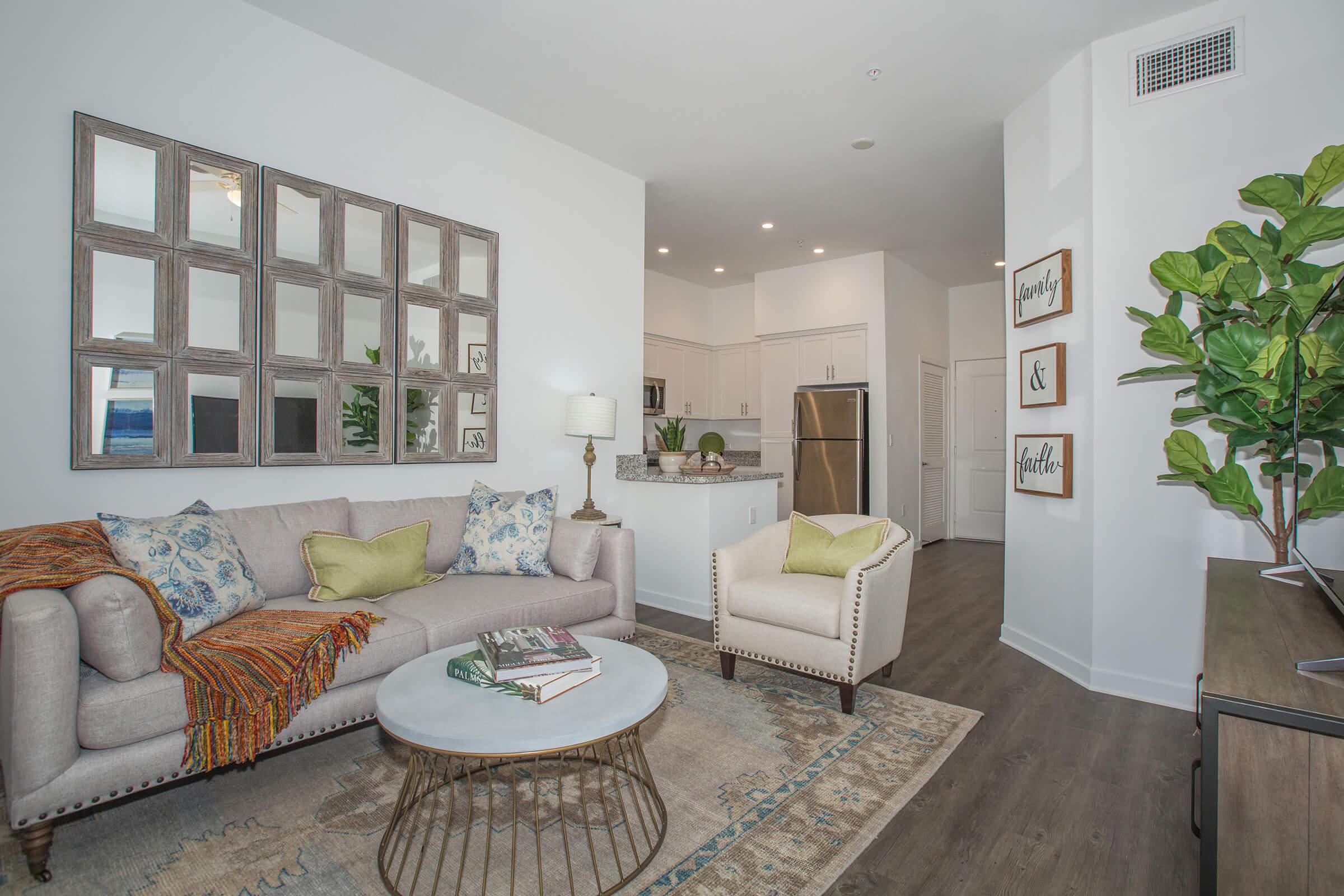 A modern living room featuring a light-colored sofa adorned with decorative pillows, a stylish coffee table, and a round area rug. A large decorative mirror hangs on the wall, and a potted plant adds a touch of greenery. The adjacent kitchen is visible, showcasing white cabinetry and stainless steel appliances.