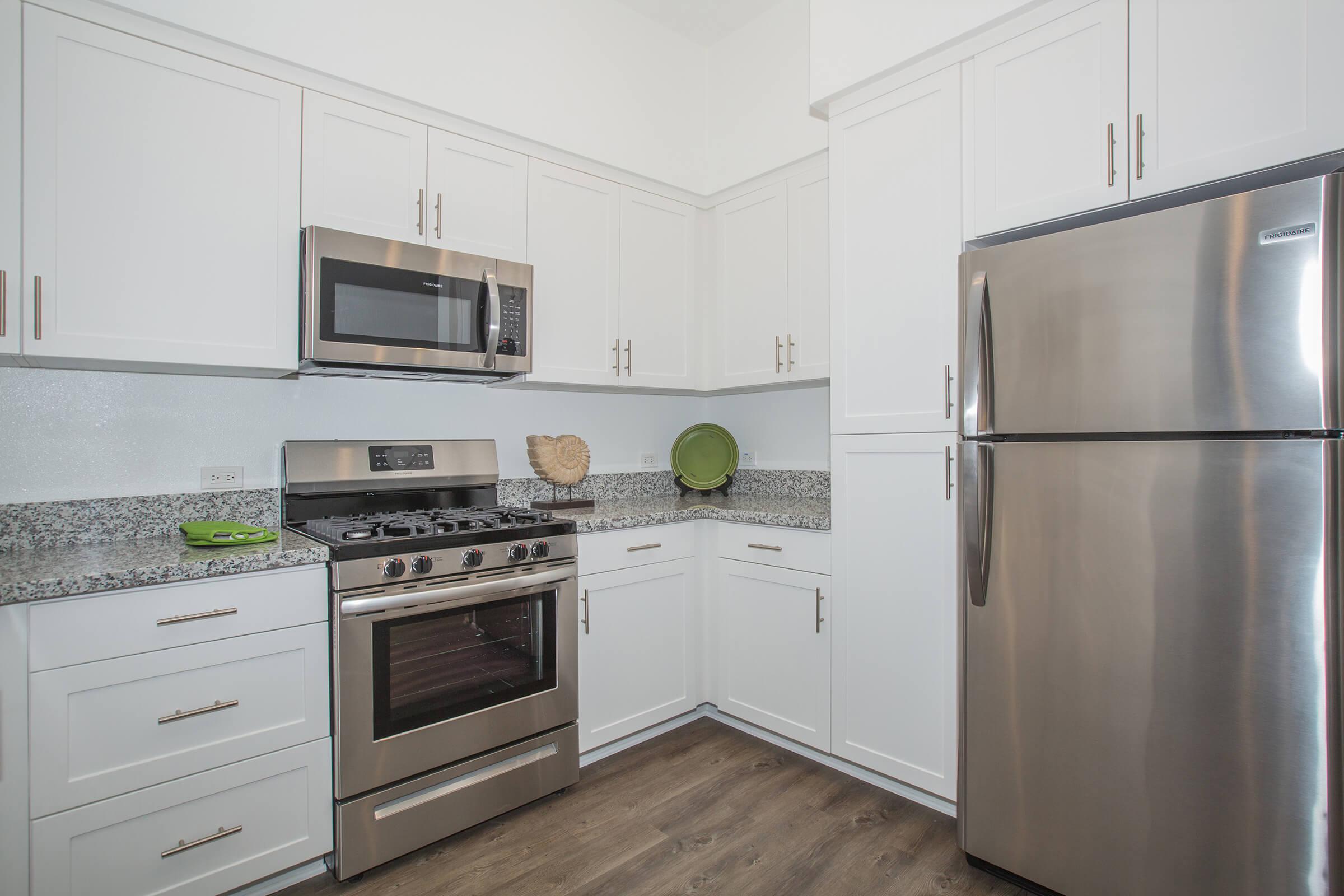 Modern kitchen featuring white cabinets, a stainless steel refrigerator, a gas stove with an oven, and a built-in microwave. The countertops are made of gray granite, and there are decorative green items on the counter. The flooring is a warm wood finish, giving the space a contemporary look.
