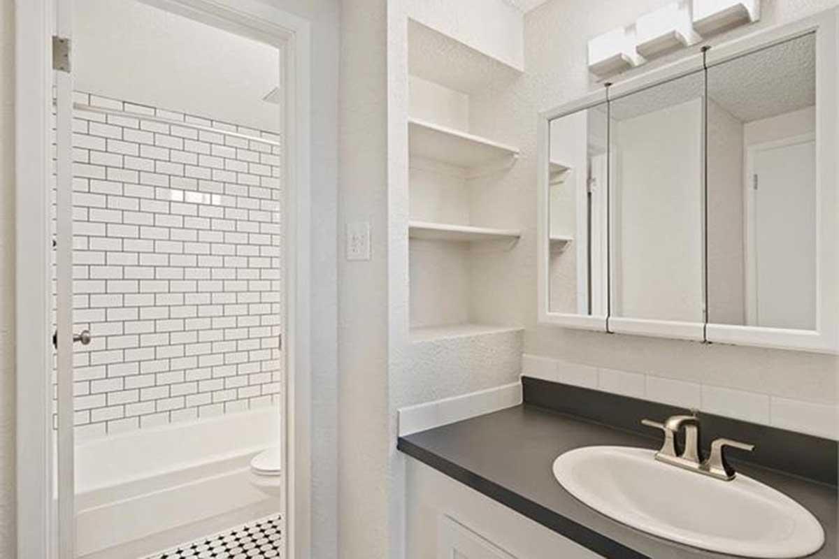 A modern bathroom featuring a white tiled shower area with a bathtub, a black countertop sink with two faucets, a wall-mounted mirror with lights above, and open shelving. The walls are painted in a light color, creating a bright and clean look. A door leads to the shower area.