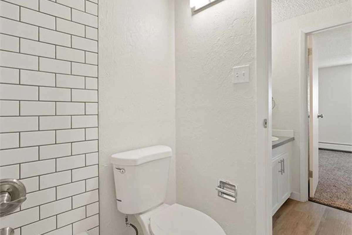 A clean, modern bathroom featuring a white toilet and a light-colored wall with white subway tiles. There is a vanity with a countertop partially visible to the left, and a doorway leads to another room in the background. The flooring is a light wood finish, contributing to a bright and airy atmosphere.