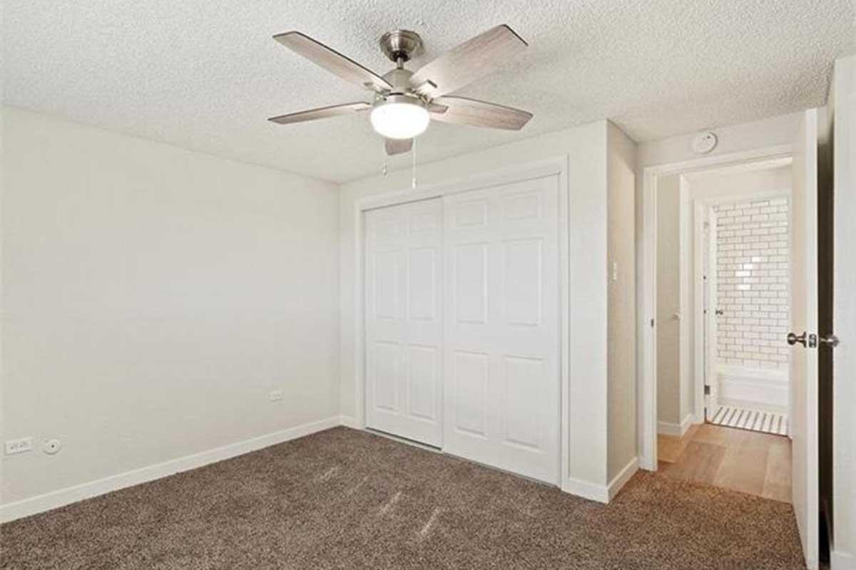 A well-lit, empty room featuring light-colored walls and carpeted flooring. The ceiling has a modern ceiling fan, and there are white double doors leading to a closet. A doorway on the right leads to a bathroom with a tiled floor visible in the background.