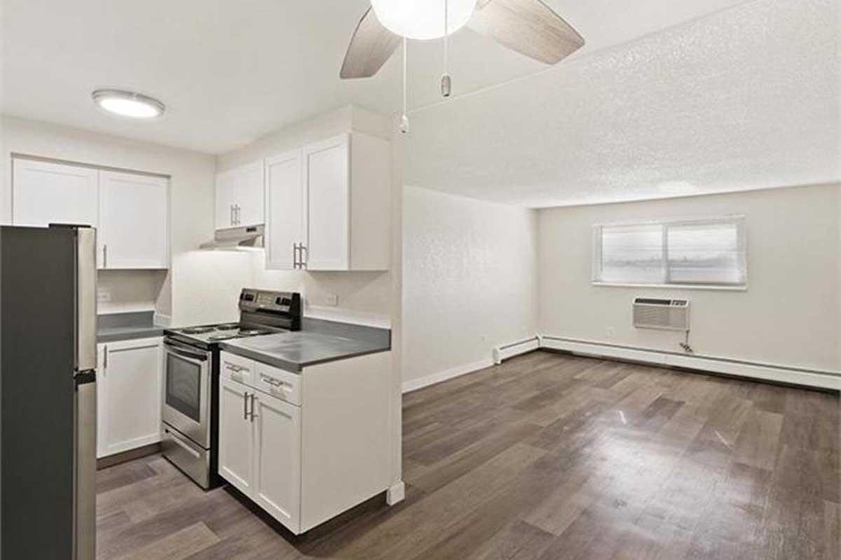 A modern kitchen with stainless steel appliances, including a stove and fridge, and white cabinetry. The adjacent living area features hardwood flooring, a ceiling fan, and a window with a view. There is an air conditioning unit on the wall, adding to the contemporary feel of the space.