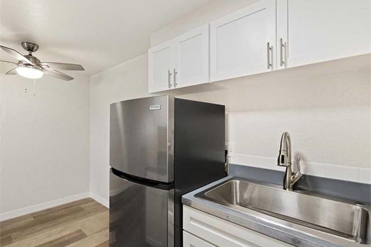 Modern kitchen with stainless steel appliances, including a refrigerator and a sink with a sleek faucet. White cabinets above the countertop and a ceiling fan add a contemporary touch. The floor features light-colored wood, enhancing the bright and airy atmosphere.