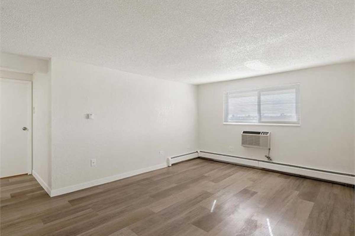 Bright, empty room with light-colored walls and wooden flooring. A window on one wall allows natural light to enter. An air conditioning unit is installed below the window. The space has a clean, minimalistic appearance, suitable for various furniture layouts.