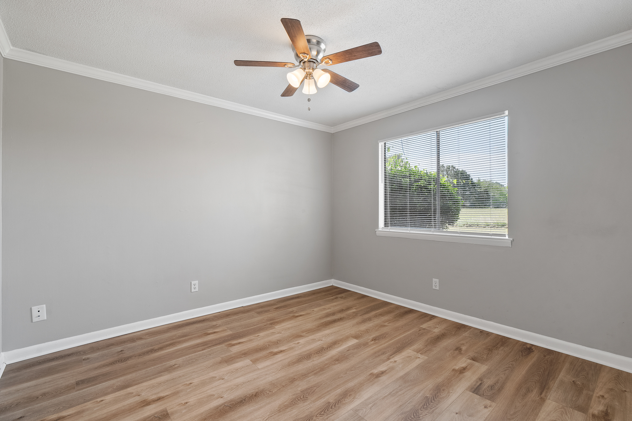 A vacant room with light gray walls, a ceiling fan with five wooden blades, and a large window letting in natural light. The floor features light hardwood laminate. There are no furnishings, creating a spacious and airy atmosphere with a view of greenery outside.