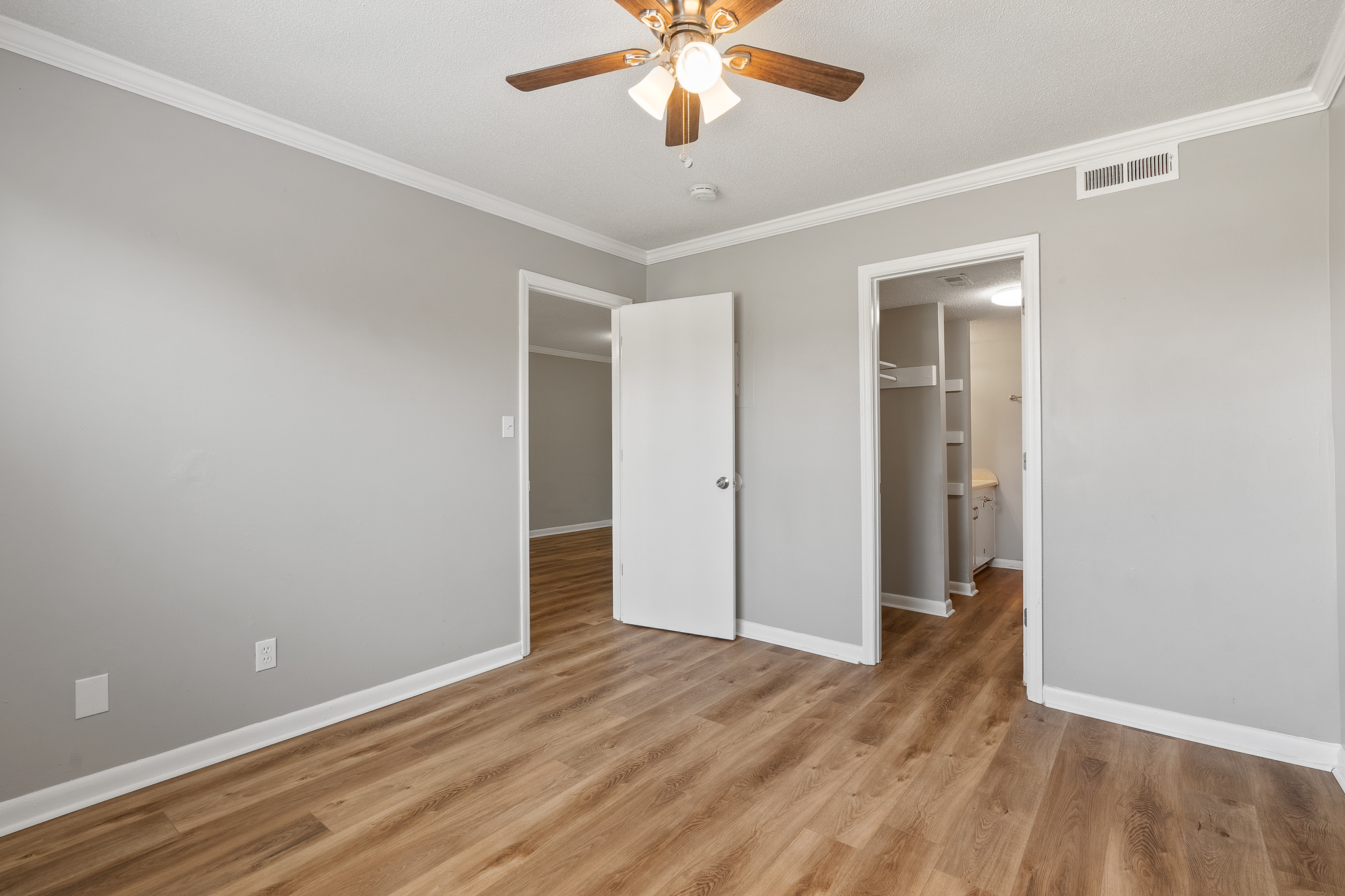 A light, airy room with gray walls and wooden flooring. A ceiling fan is visible, and there are two doorways—one leading to a small bathroom area and another to an adjoining room. The room is empty, creating a spacious feel.