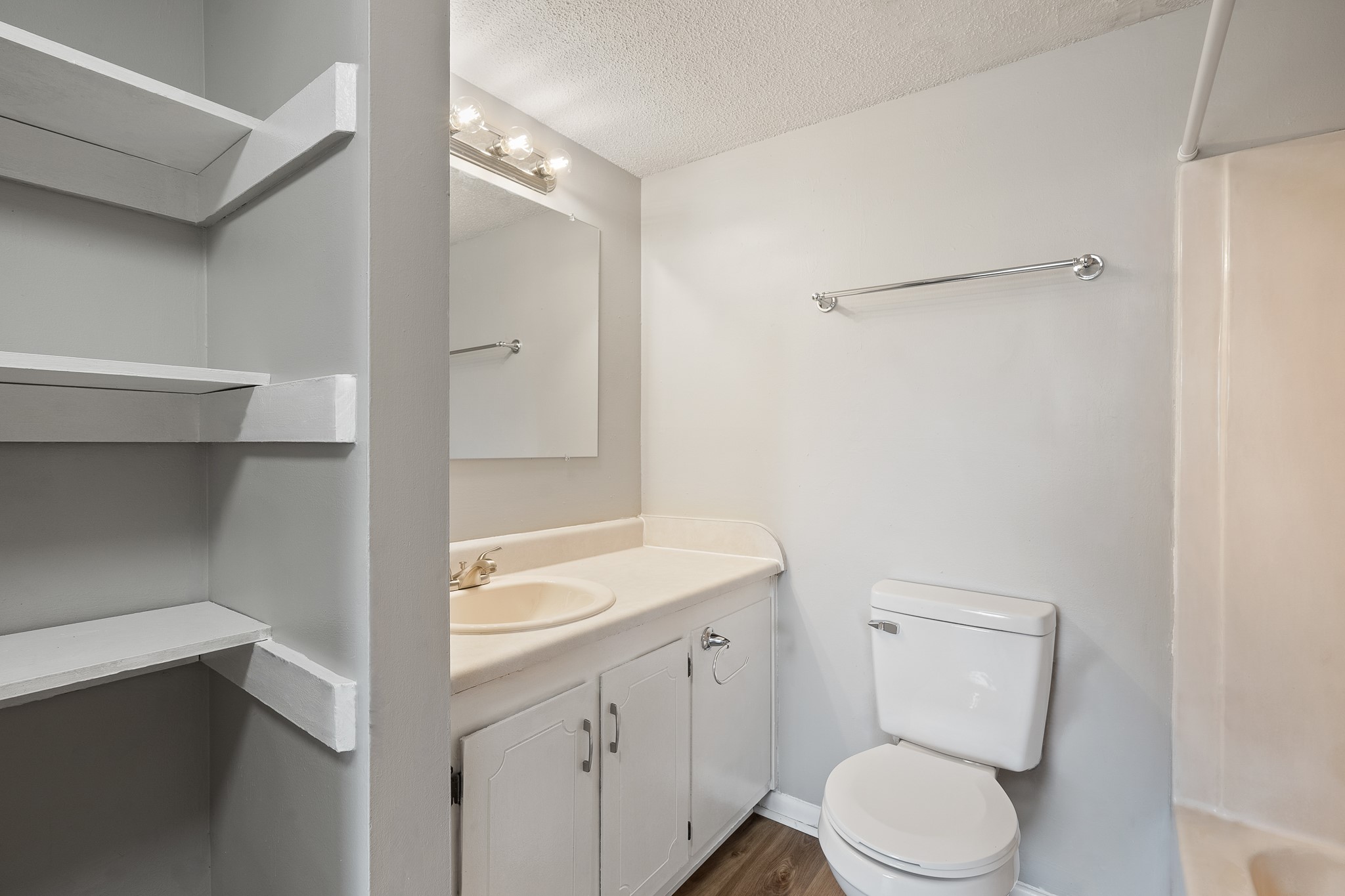 A clean, well-lit bathroom featuring a white toilet, a sink with a light-colored countertop, a large mirror above the sink, and a towel rack. There are open shelves on the left side, and a shower area is visible on the right. The walls are painted light gray, contributing to a spacious and modern appearance.