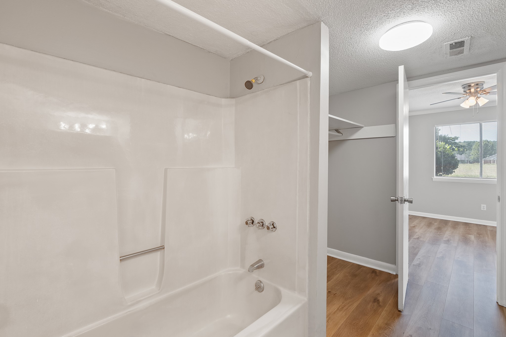 A clean and modern bathroom featuring a white bathtub with a showerhead, light gray walls, and wooden flooring. An open doorway leads to a well-lit room with a ceiling fan and a window overlooking greenery outside. Natural light enhances the spacious feel of the area.