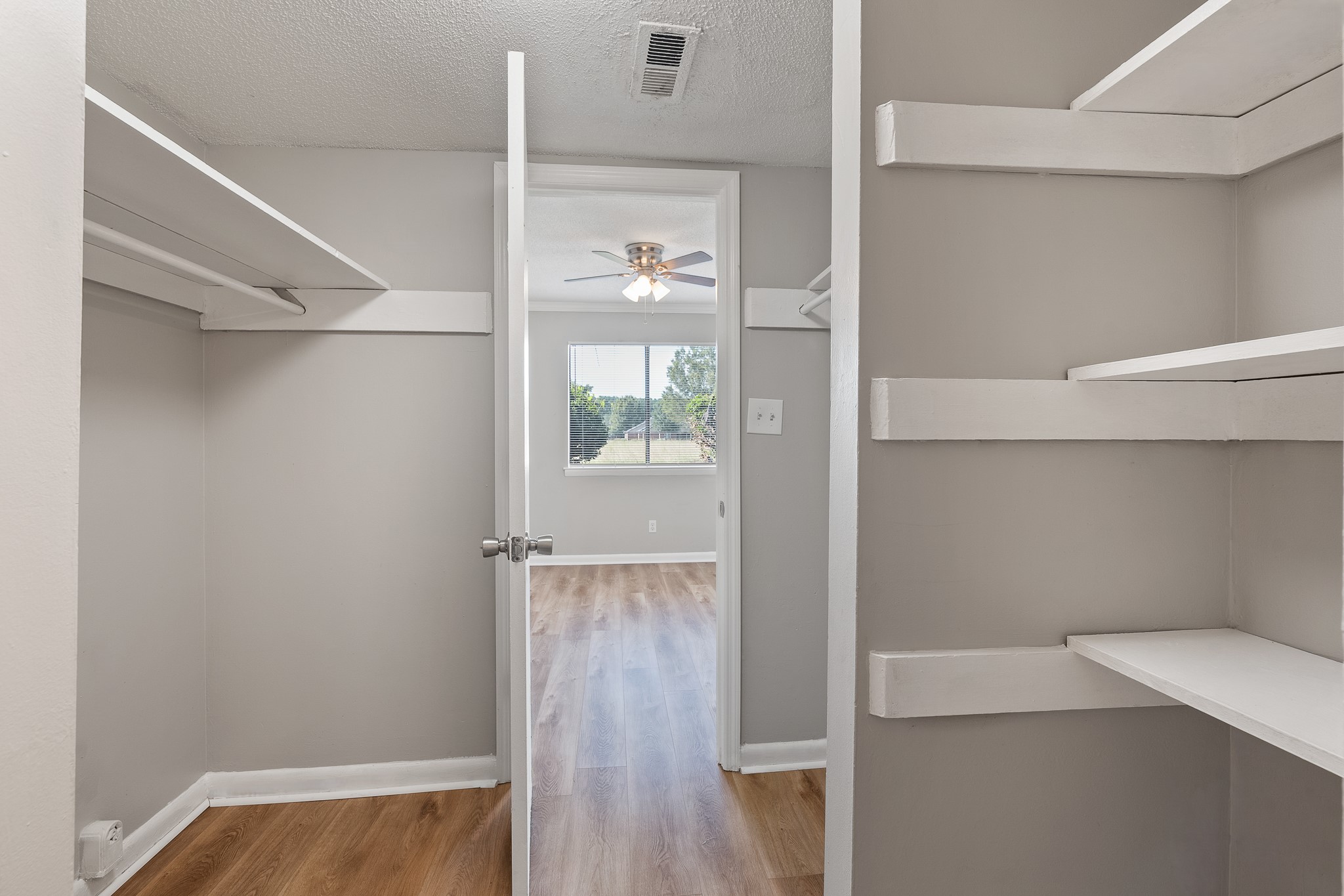 A view of a light-colored room with a ceiling fan, showcasing an open closet space with white shelving. The room features a window allowing natural light, and the floor has a wooden finish. The walls are painted in a neutral tone, creating a bright and spacious atmosphere.