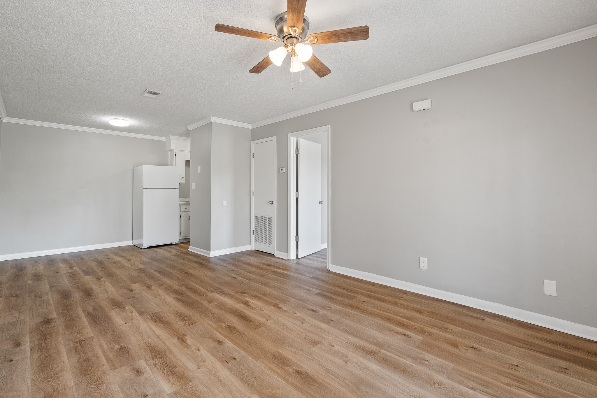 A spacious, unfurnished living area with light gray walls and wooden flooring. A ceiling fan with wooden blades hangs above. To the left, a kitchen area with a white refrigerator is visible. A doorway on the right leads to another room or hallway. Natural light brightens the space, creating an inviting atmosphere.