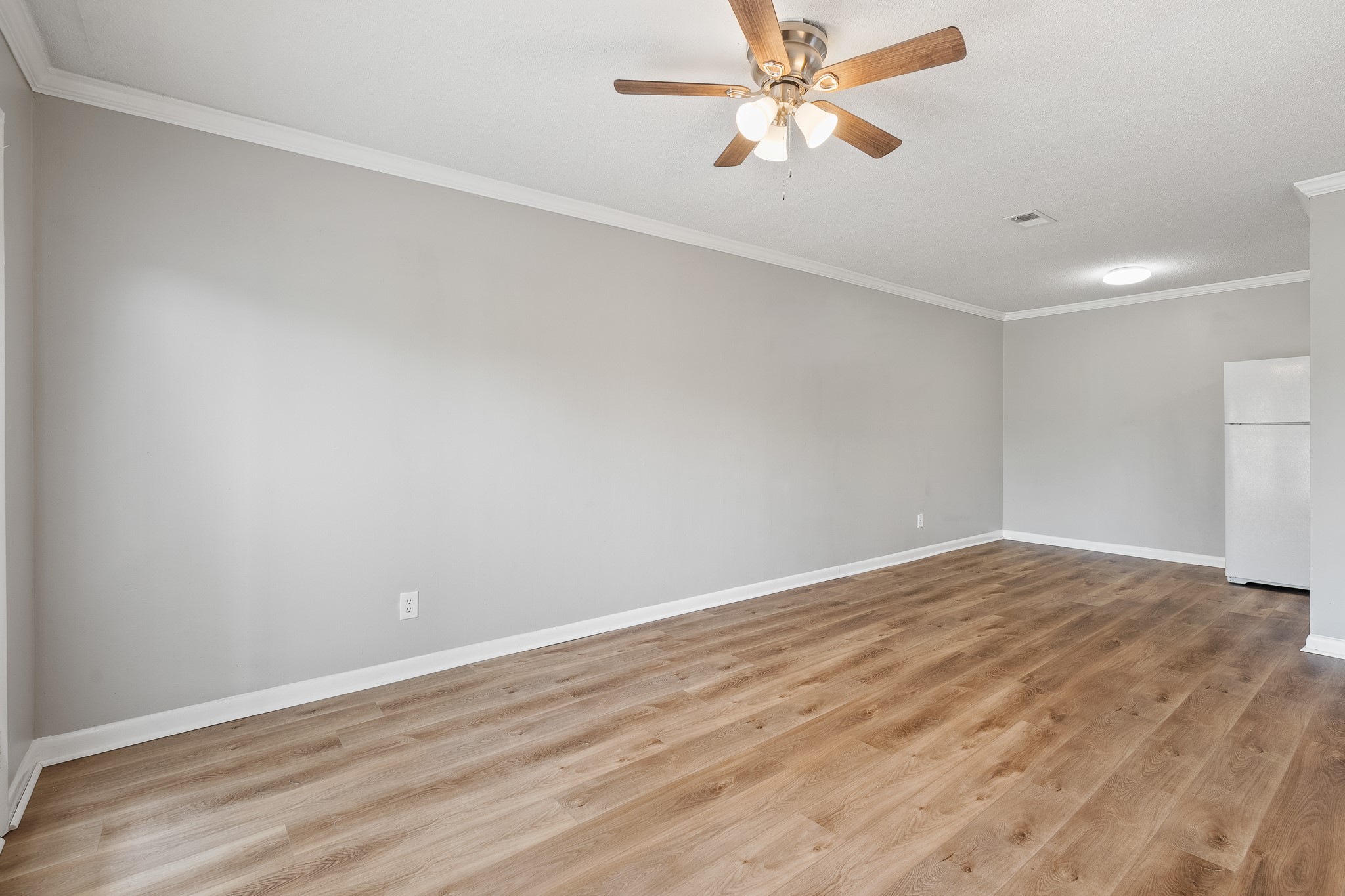 A spacious, well-lit living room featuring light gray walls, hardwood flooring, and a ceiling fan with three lights. In the background, a white refrigerator is visible against a wall in an adjacent area, contributing to the open layout design of the space.