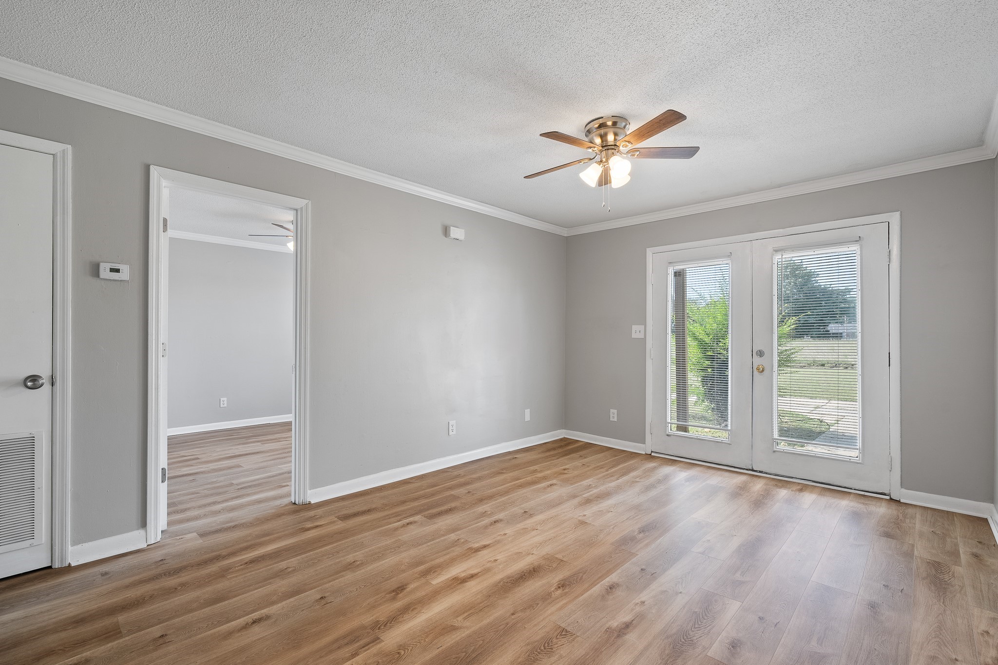 A bright, unfurnished living room with light wood flooring. The room features a ceiling fan and large glass doors leading to an outdoor area. The walls are painted in a soft gray, and there is a door on the left leading to another space. Natural light floods the room, creating a warm and inviting atmosphere.