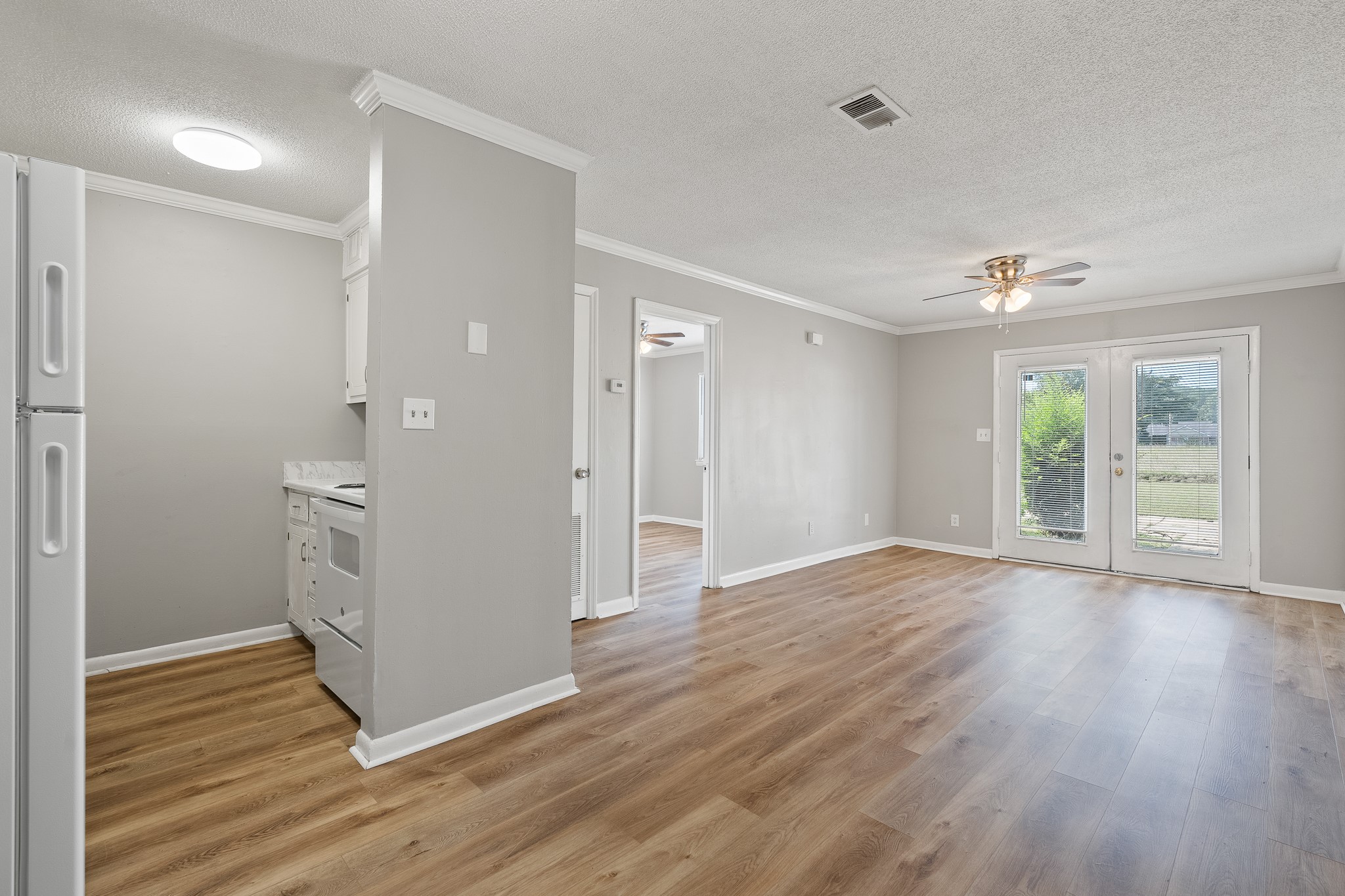 A spacious, empty living area with light grey walls and laminate wood flooring. On the left, a white refrigerator and kitchen area are visible, while the right features a set of sliding glass doors leading to an outdoor view. Ceiling fan light provides illumination in the room, enhancing the open feel.