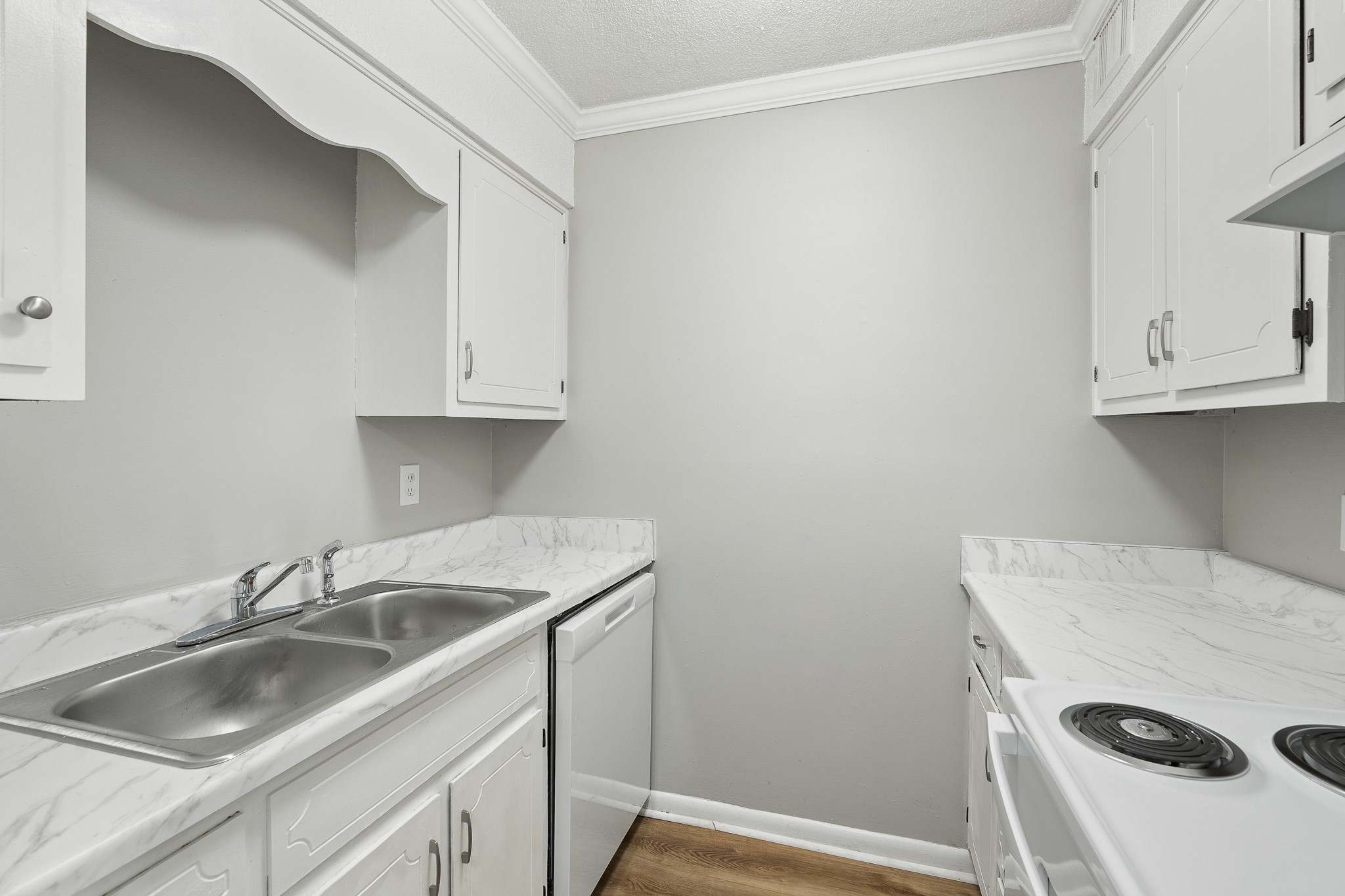 A compact kitchen featuring white cabinets, a double sink, and a marble-look countertop. The space includes a white dishwasher and an electric stove with two burners. The walls are painted light gray, and the floor has a wood-like finish, creating a clean and modern atmosphere.