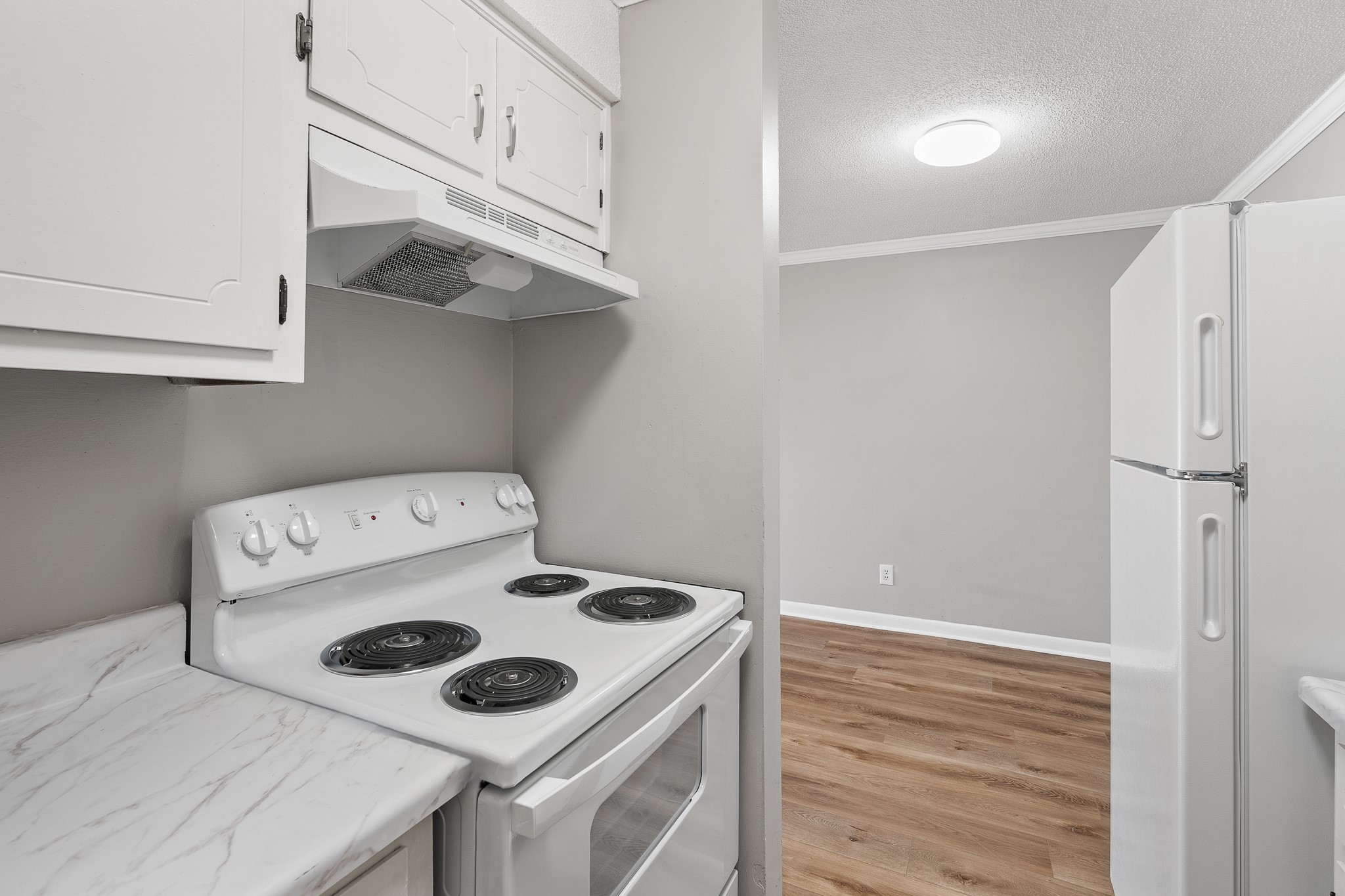 A modern kitchen featuring a white electric stove with four burners, a range hood, and light-colored cabinets. The countertop is a marble pattern, and a white refrigerator is visible in the background. The walls are painted in a soft gray, and the floor is comprised of light wood planks, creating a bright and inviting space.